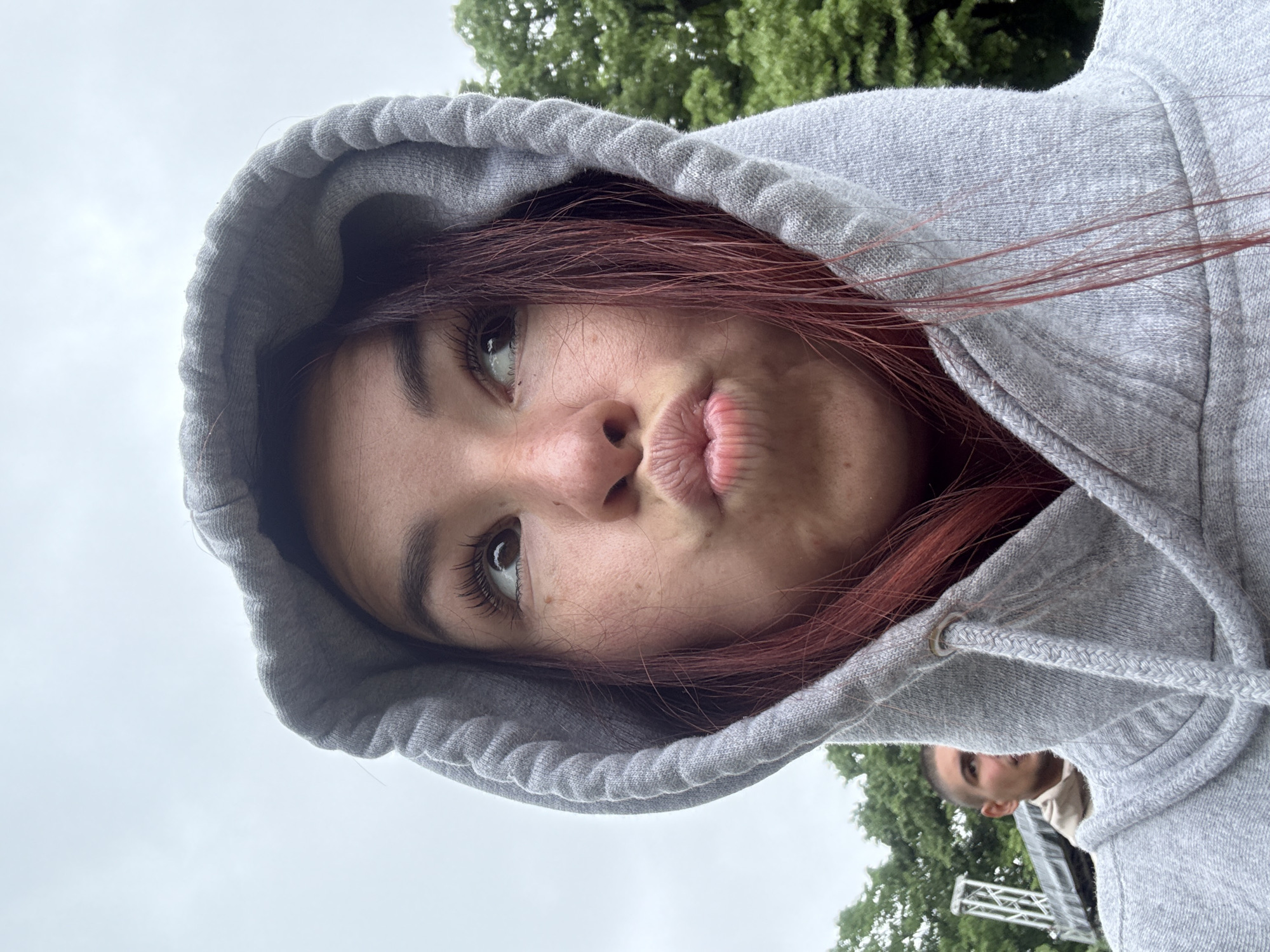 A selfie shows a person with fair skin, brown eyes, and reddish-brown hair wearing a gray hooded sweatshirt. The hood is up, framing the person's face, which is posed with pursed lips. The person is looking slightly upward and to the left. Part of another person's head is visible in the lower right corner of the frame, along with a structure that appears to be scaffolding. The background includes green trees and a pale sky. The specific location in Kozloduy, Bulgaria, cannot be determined from the image.