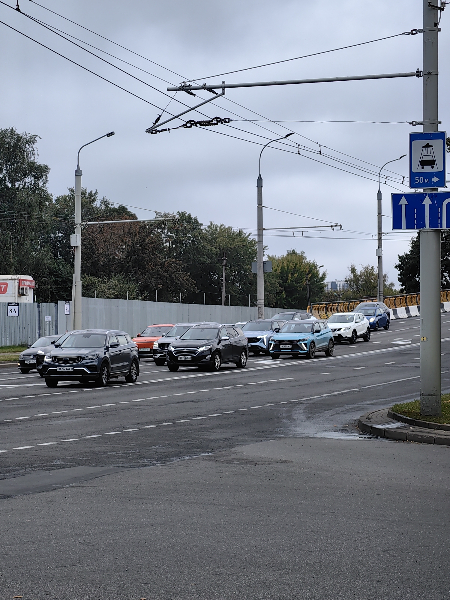 The image depicts a traffic scene on an overcast day in Minsk, Belarus. A line of cars is moving along a multi-lane road. In the foreground, a dark grey SUV is prominent, followed by a lighter grey SUV and several other vehicles of various colors and types. The road surface appears wet, with visible lane markings. Overhead, a complex network of trolleybus wires is suspended by poles and cables. To the right of the frame, traffic signs are visible, including one indicating a car wash 50 meters ahead and another with directional arrows. In the background, trees line the street, and a chain-link fence with an adjacent corrugated metal fence is present. A light-colored building with some signage is partially visible behind the fence. The overall impression is of a typical urban street scene in a moderately busy city.