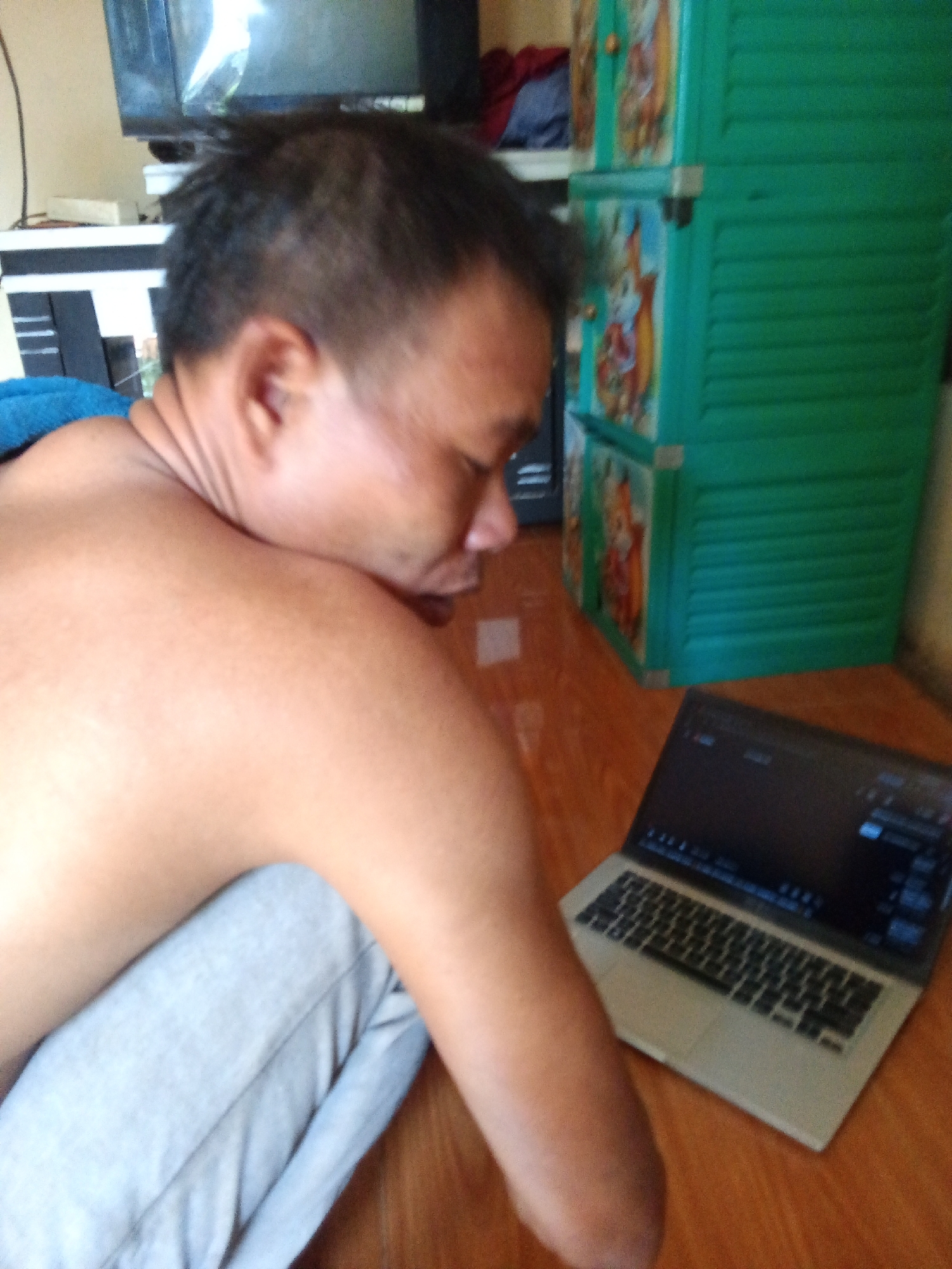 The image shows a man in Subang, Indonesia, from the shoulders up, viewed from behind and to the side. He is leaning his head down, facing a laptop that is open on a wooden floor. The man is wearing light-colored pants. In the background, a green plastic cabinet with cartoon images on its doors is visible, as well as a television set on a stand. The lighting suggests it is daytime, with natural light coming from the left.