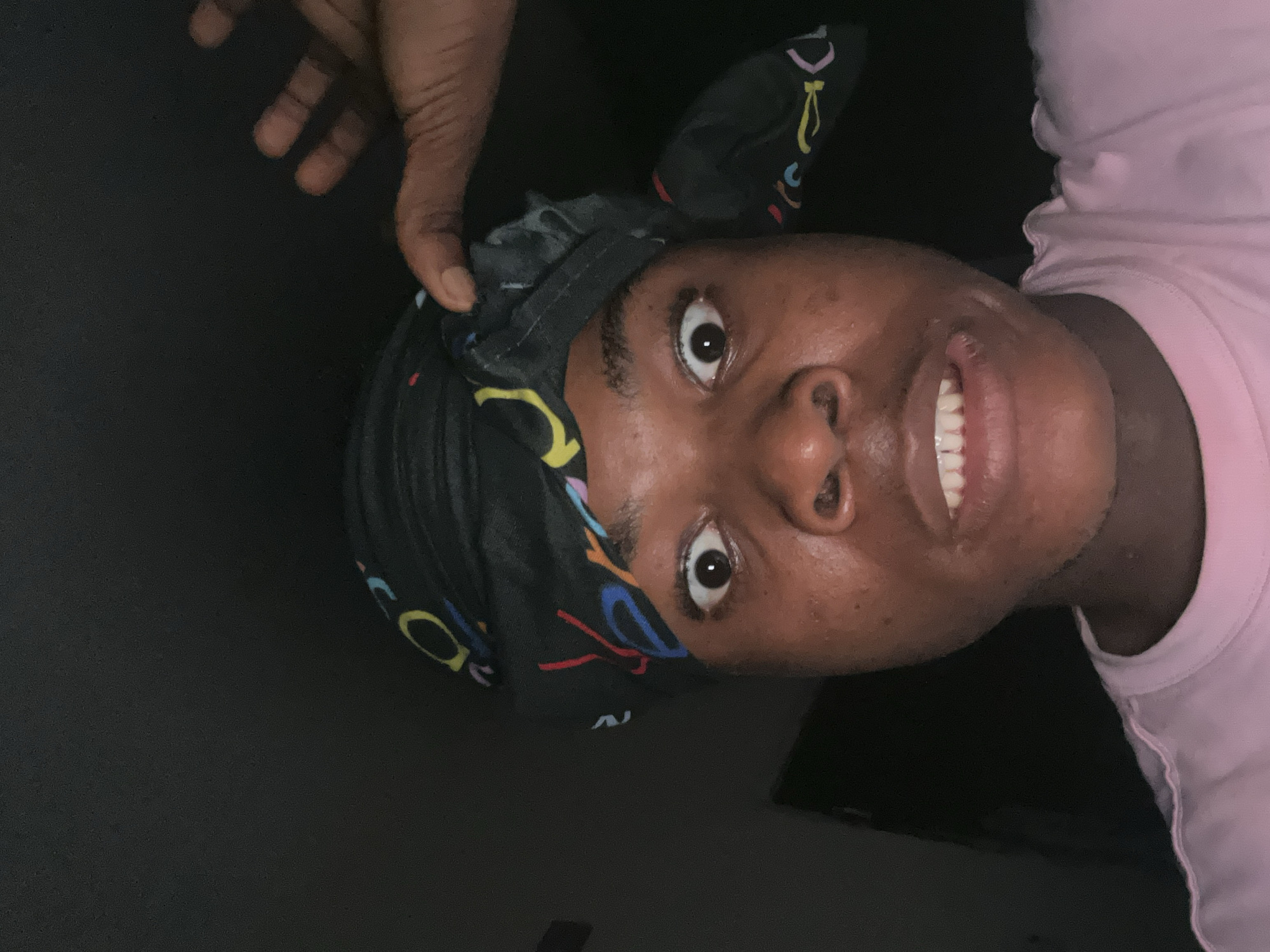 A close-up, front-facing, low-angle photograph captures a Black individual wearing a black bandana with colorful abstract designs. The individual's eyes are wide open, and their mouth is slightly agape, revealing their teeth. The background is dark and indistinct, suggesting an indoor setting at night. A hand, also Black, is visible in the upper right corner, lightly touching the bandana on the individual's head. The individual is wearing a light pink t-shirt. The scene appears to be a selfie taken in Ihiagwa, Nigeria.