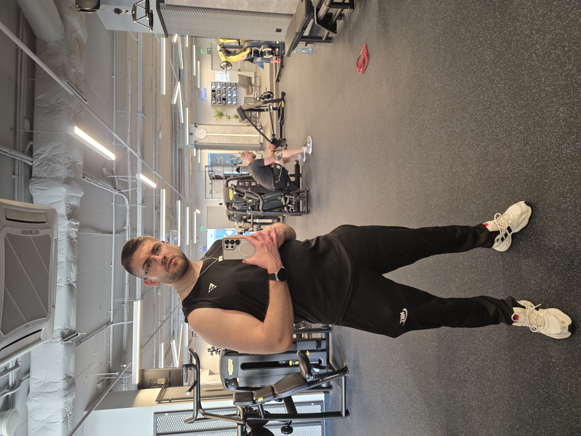 A man is standing in a gym, facing the camera, holding a smartphone in his hands as if taking a selfie. He is wearing a black sleeveless shirt and black Nike sweatpants, and white sneakers. A black watch is on his left wrist. The gym appears to be well-lit with artificial lighting and features various exercise machines. Another person is using a machine in the background. The location is likely a gym in Plovdiv, Bulgaria, but there is no direct evidence of its location.