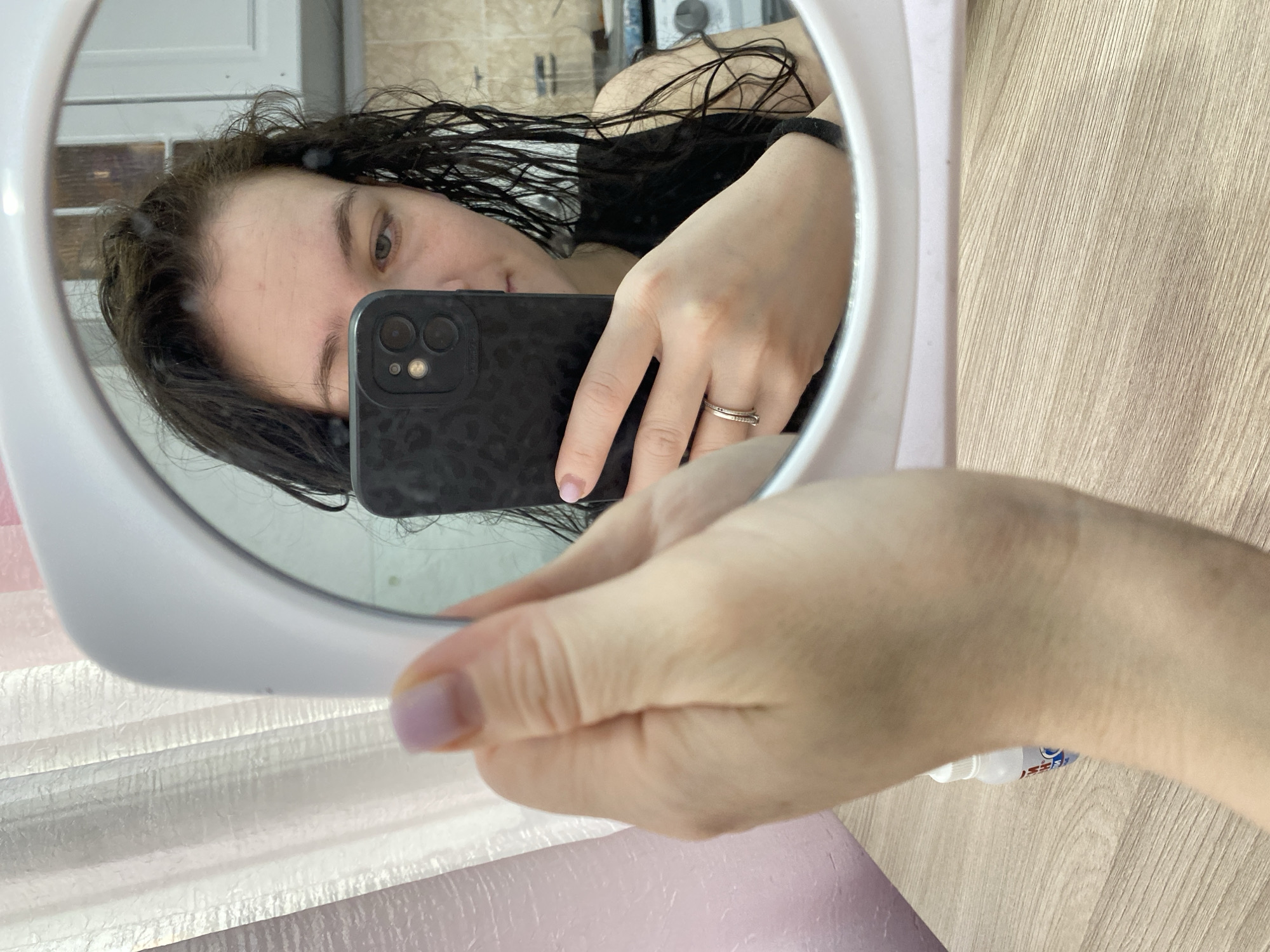 A woman is taking a selfie in a round mirror. The woman has dark, wet hair. She is holding a dark-colored smartphone in her right hand and is wearing a ring on her finger. The bathroom appears to have light wood-grained surfaces and a pink striped surface near the bottom of the frame. There is a white object near the bottom right of the frame, possibly a bottle. The location is inferred as a domestic bathroom but there is no visual indication that this is in the city of Chaikovsky, Russia.