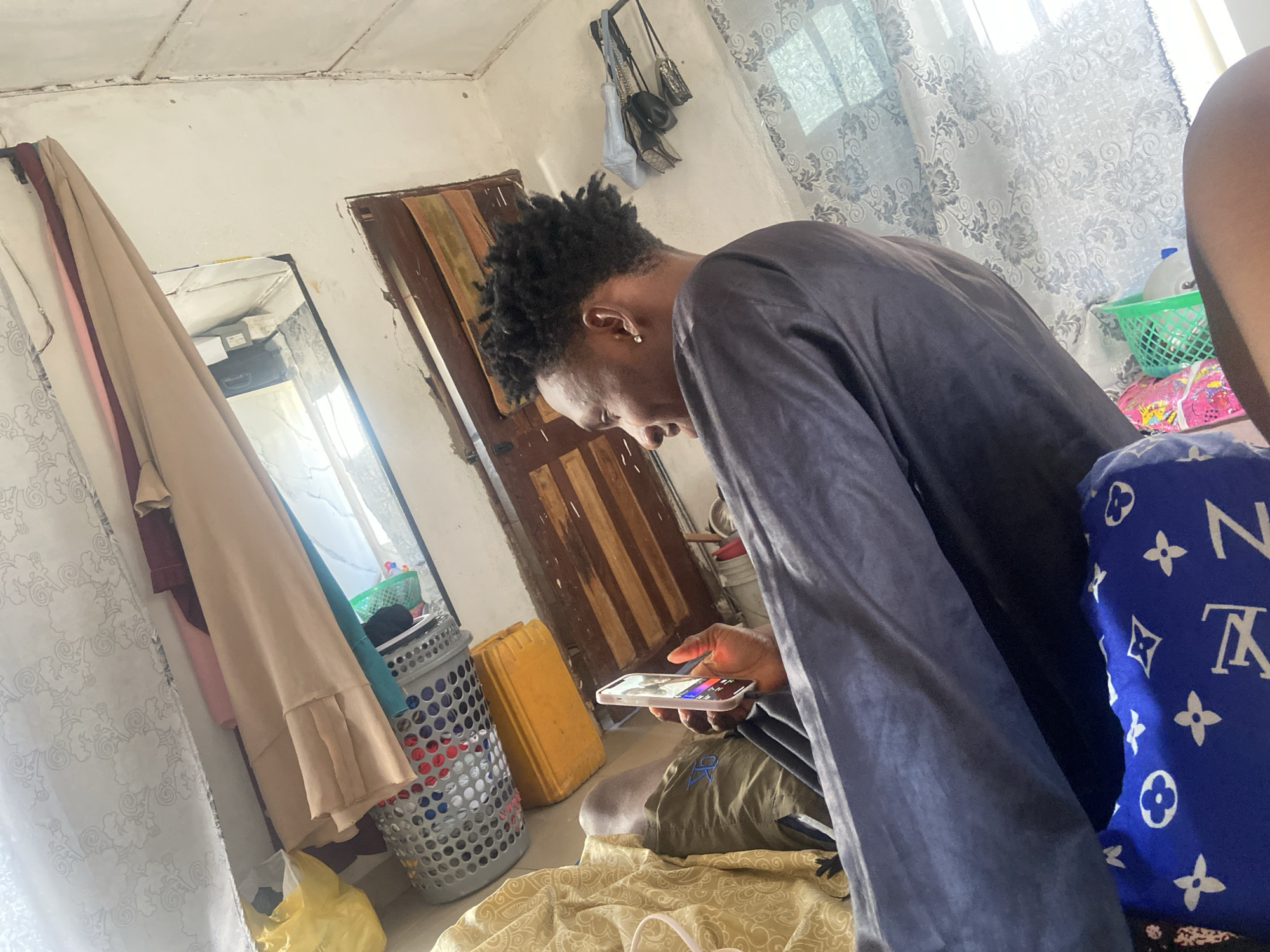 A young Black man with short, dark, textured hair and a small earring is using a smartphone. He is wearing a long-sleeved gray shirt and olive green shorts. The man is sitting on the floor of a room that appears to be inside a home in Lokoja, Nigeria. The environment includes a mirror with clothing hanging nearby, a door with a wooden panel design, a laundry basket, and a yellow container. A blue pillow with the Louis Vuitton logo is visible next to him. Curtains adorn the window.