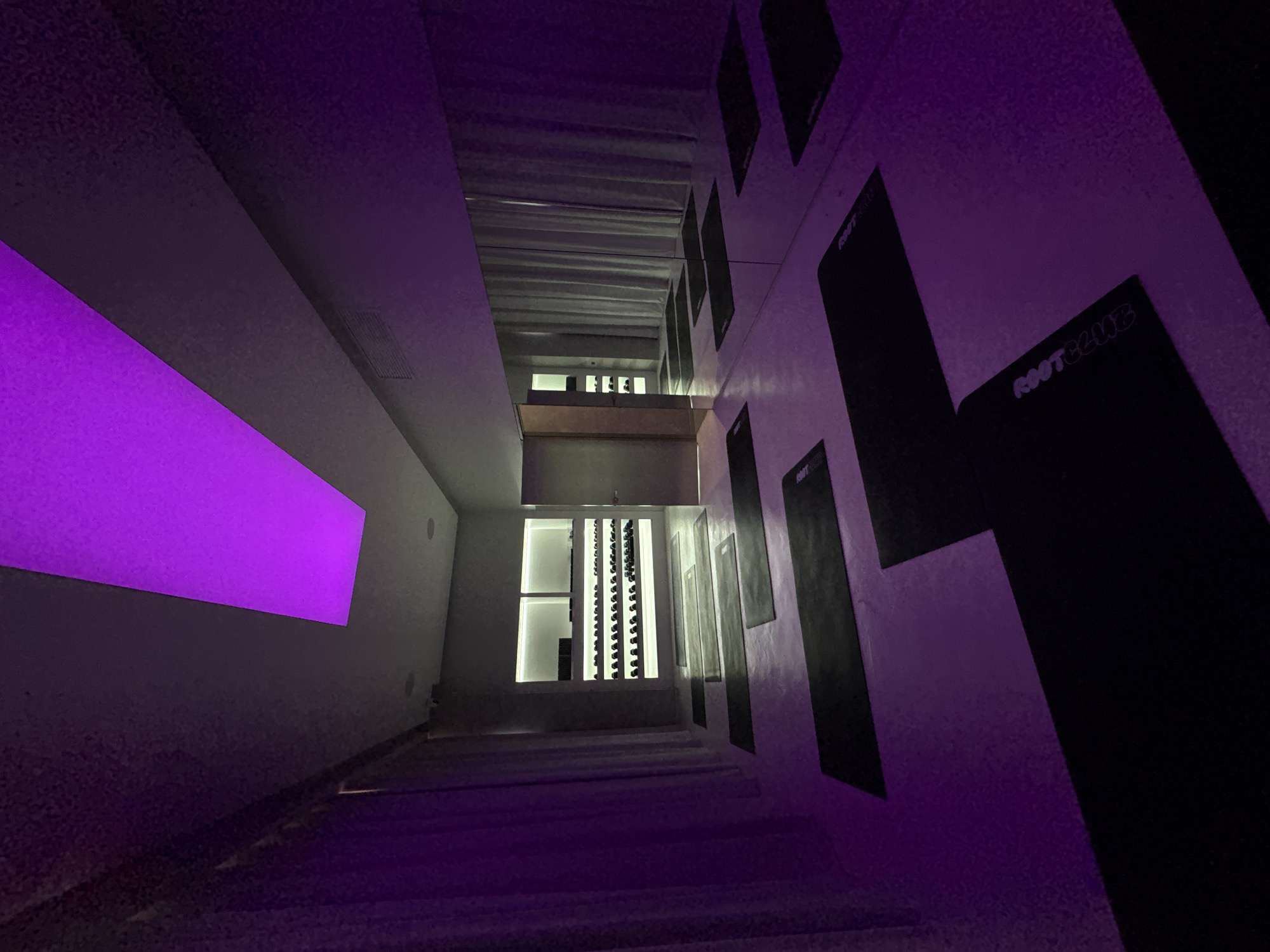 An interior view of a dimly lit yoga studio in Mexico City, Mexico. The space is illuminated by a large rectangular purple light fixture on the ceiling and purple ambient lighting. Black yoga mats are neatly arranged on the floor in a grid pattern. To the left, a floor-to-ceiling curtain is drawn. In the background, a white shelving unit displays rows of water bottles and other items. A doorway is visible to the right of the shelves, leading to another room. A large mirror covers a significant portion of the right wall, reflecting the yoga mats and curtains. There are no people present in the image.