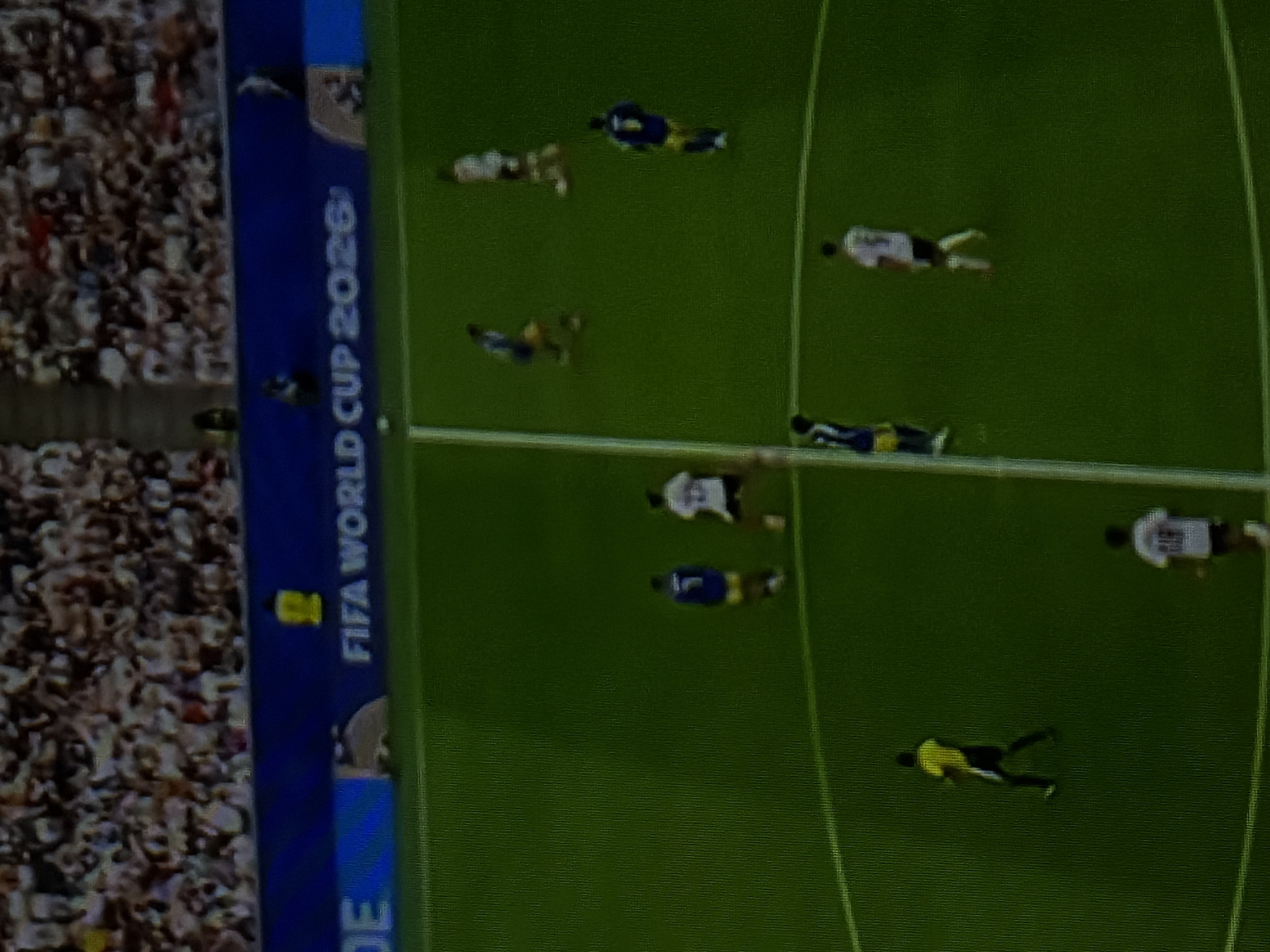 An aerial, full-frame, high-angle shot captures a soccer game. The stadium is filled with a blurry crowd of spectators. A banner that reads "FIFA WORLD CUP 2026" is visible at the top of the frame. The soccer field is green with white lines. Several players, wearing yellow, blue, and white jerseys, are scattered across the field. One player in a yellow jersey is standing near the left sideline, looking towards the right. Another player in a white jersey is standing in the lower right quadrant of the frame. Two players in blue jerseys and one player in a white jersey are in the upper right quadrant, with one player in blue appearing to be kicking a ball. Another player in a yellow and blue jersey is in the center left of the frame. The game appears to be in progress.