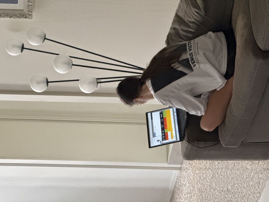 An indoor scene within a residential dwelling in Venice, Italy. A female individual is positioned with her back to the viewer, seated cross-legged on a grey fabric sofa. Her dark hair is secured in a low ponytail. She wears a white long-sleeved shirt adorned with a black graphic on the back displaying the text "VENUS VICTRIX," and dark shorts. She is actively operating an open black laptop computer, which rests on her lap. The laptop screen shows a color-coded spreadsheet or data table with red, orange, and green sections; a charging cable is connected. To the right of the individual stands a modern floor lamp featuring five black stalks, each topped with a spherical white opaque globe. The surrounding walls are light-colored. To the left, a framed doorway leads into an adjacent room. The visible flooring throughout the space is terrazzo, an architectural feature consistent with Italian residential design.