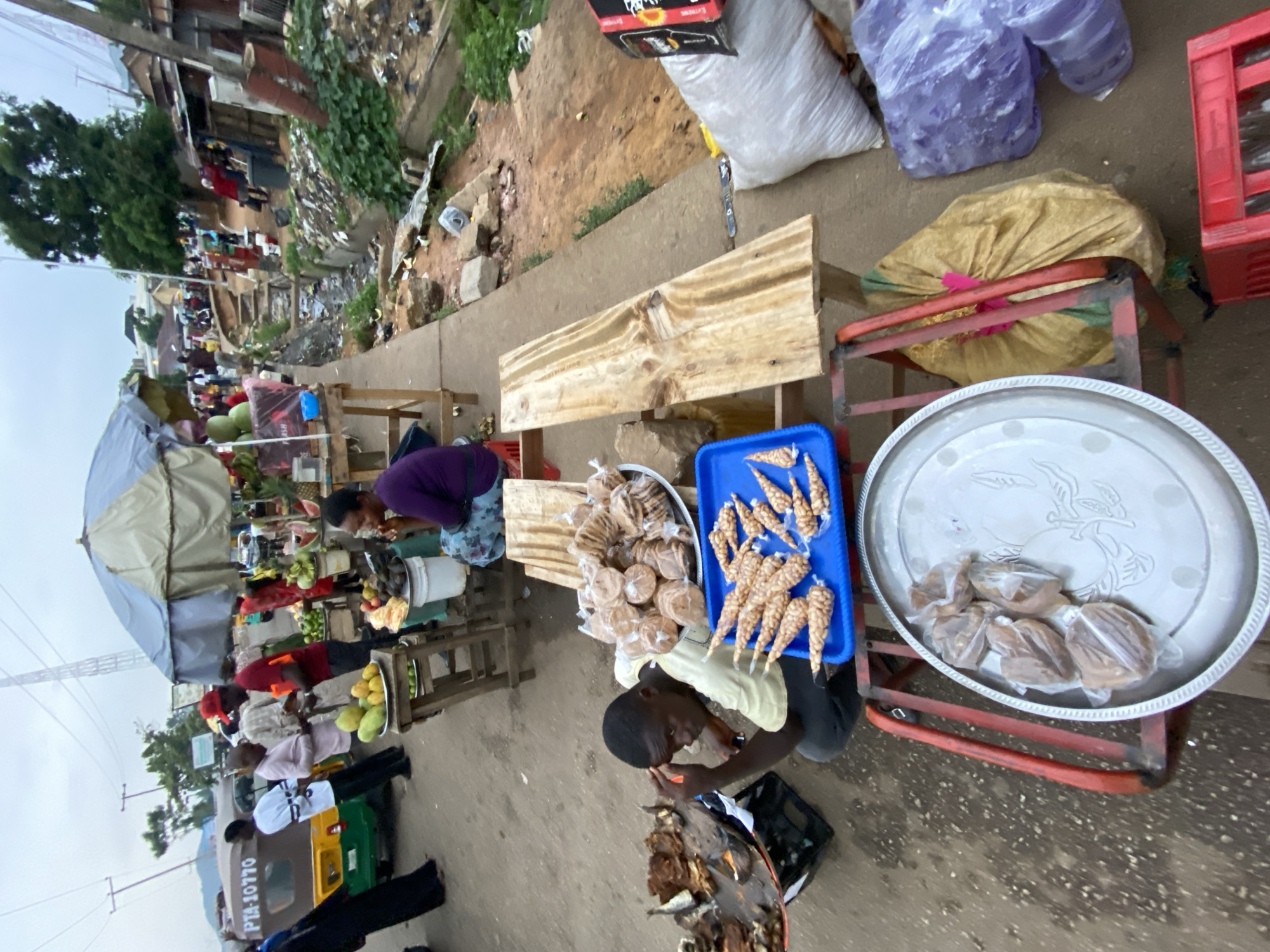 The image depicts a street scene in Bukuru, Nigeria, showcasing informal vending activities. Several vendors are present, one of whom is attending to a selection of goods that include individually wrapped food items displayed on trays and in a blue plastic container. Other vendors are selling fruits under a makeshift tent.
The background of the image captures a congested street with foot traffic and commercial buildings lining the sides. Evidence of urban conditions is also visible. A three-wheeled vehicle is parked near the vendors, partially visible at the lower left.