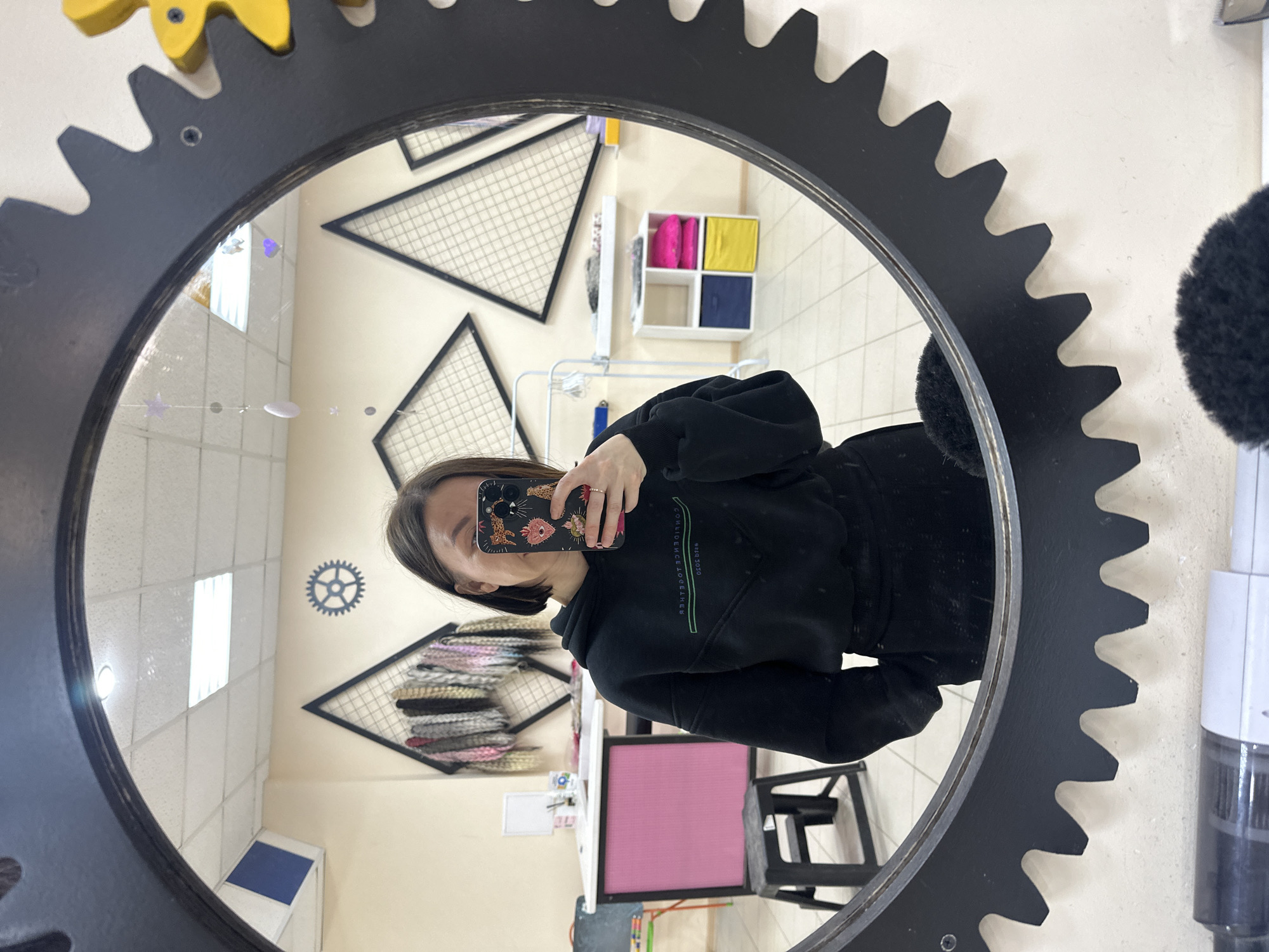 A person is taking a selfie in a mirror shaped like a gear. The reflection shows a hair salon with white walls, black triangular shelves, a display of stacked hair extensions or wigs, a small shelf unit with colorful cubes, and a smaller gear-shaped wall decoration. The person is wearing a black hoodie and holding a phone with a patterned case. The mirror itself is dark grey/black with a gear-like outer rim, possibly made of metal or a hard plastic-like material, and mounted on the wall. The location might be a beauty salon or hairdressing studio. No specific information indicates this is in Votkinsk, Russia, beyond the assumption that the person taking the photograph is physically present at the location that the metadata indicates.
