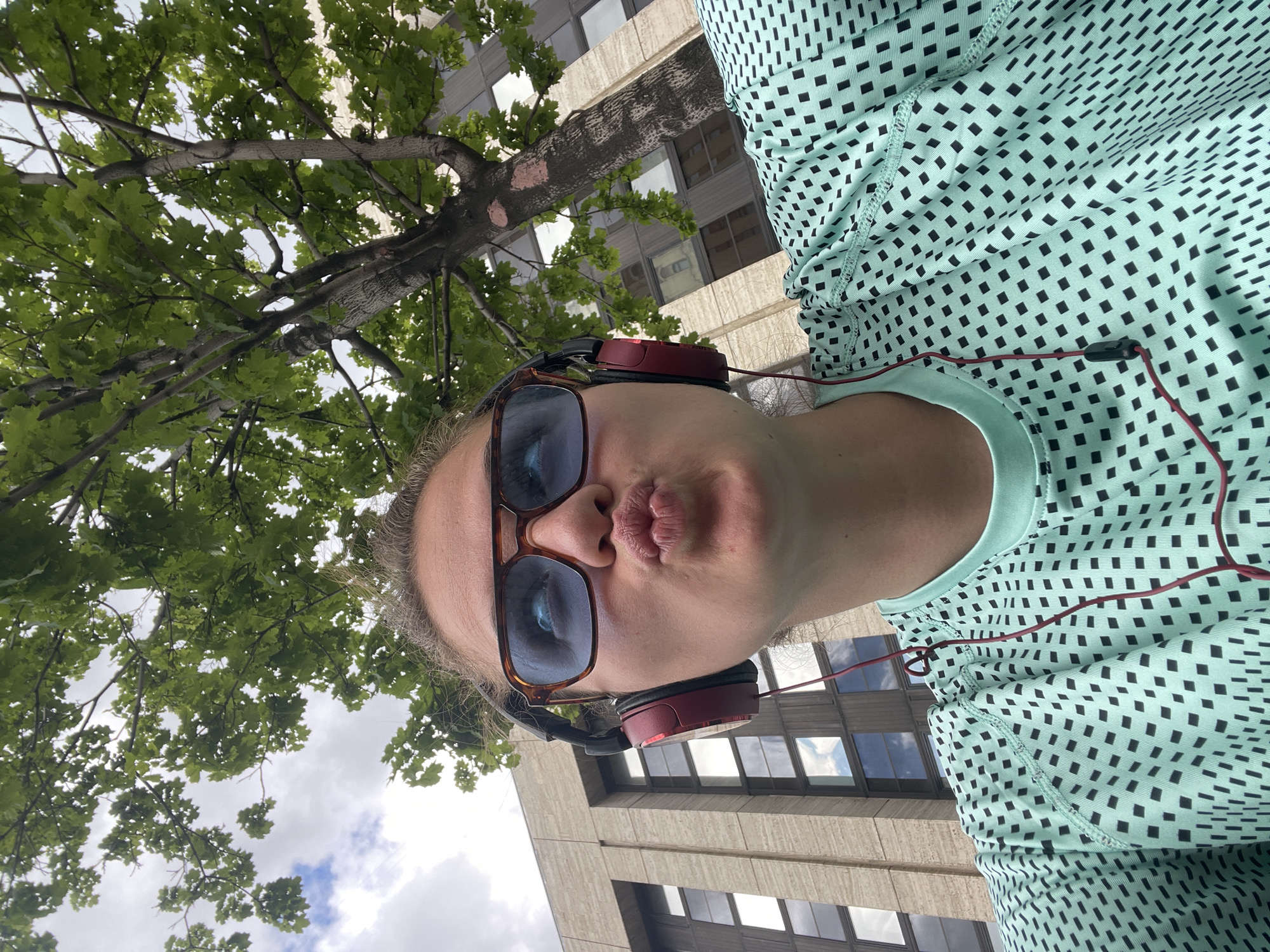 A selfie taken from a low angle looking upwards, showing a person with sunglasses and red headphones, puckering their lips. The person is wearing a light green top with small black squares. Behind them, there is a tree with green leaves and a tall, grey building with windows. Without additional information, it's impossible to verify the location as Moscow, Russia, or any other specific place. The image contains a person, architecture, and nature elements.
