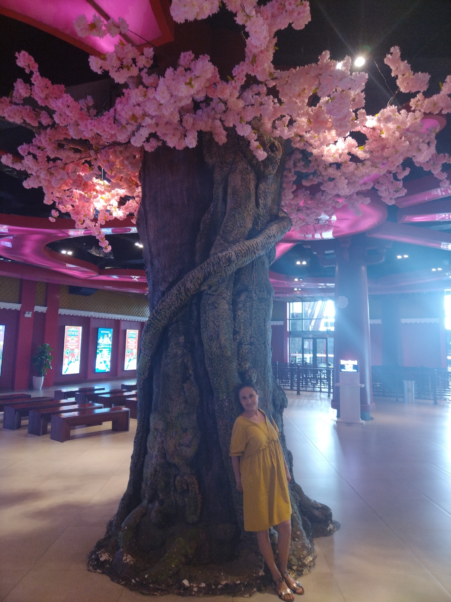 A woman in a yellow dress stands next to a large artificial tree with pink blossoms in what appears to be an indoor shopping mall or entertainment complex. The tree is centrally positioned, acting as a focal point. The background includes empty seating areas with benches, decorative posters, and large windows letting in natural light. The ceiling has pink, circular design elements and recessed lighting. Based on visual evidence and location data, this scene is located in the city of Hong Gai Ward, Vietnam.