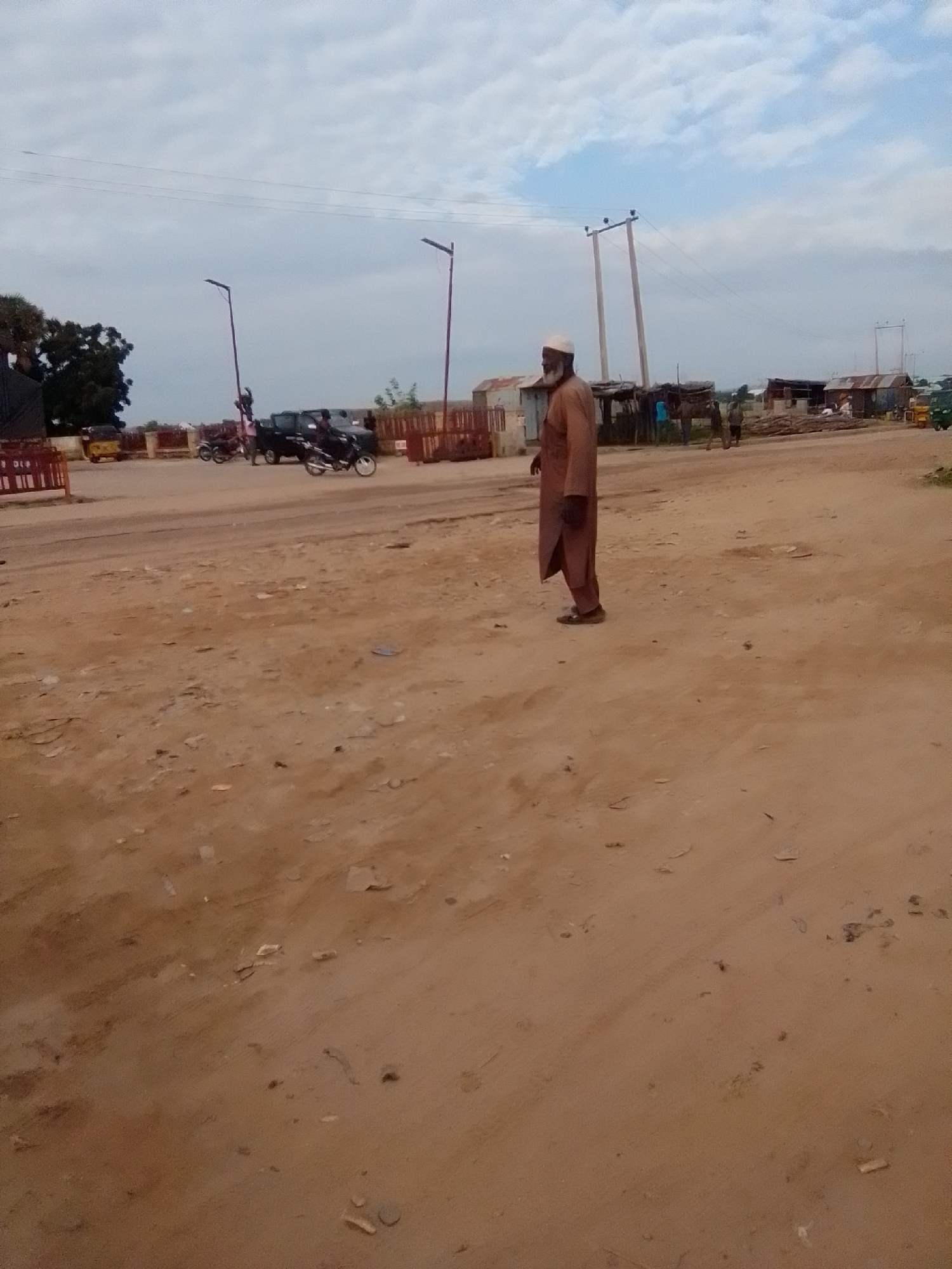 A man in a brown kaftan and cap stands on a dirt road in Gombe, Nigeria. He is facing to the right, looking at something off-camera. In the background, there are several buildings made of corrugated metal and wood, some with thatched roofs. There are also some vehicles, including a tricycle and a motorcycle. The sky is cloudy. The scene appears to be a busy market or transportation hub.