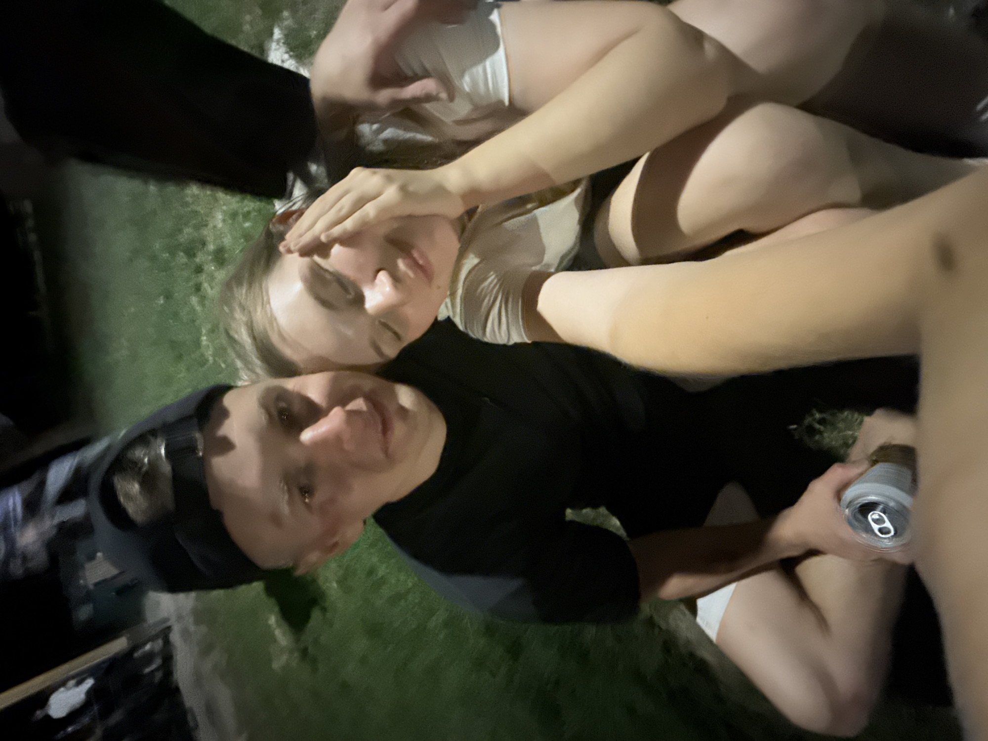 The image depicts an outdoor scene at night, possibly in a park or similar grassy area. Three individuals are visible. One individual, wearing a black shirt and cap, is holding a can and looking at the camera. Another individual is lying down with their hand covering their eyes. A third individual is partially visible. The overall environment appears dark. No specific details relate to Cukarica Urban Municipality, Serbia.