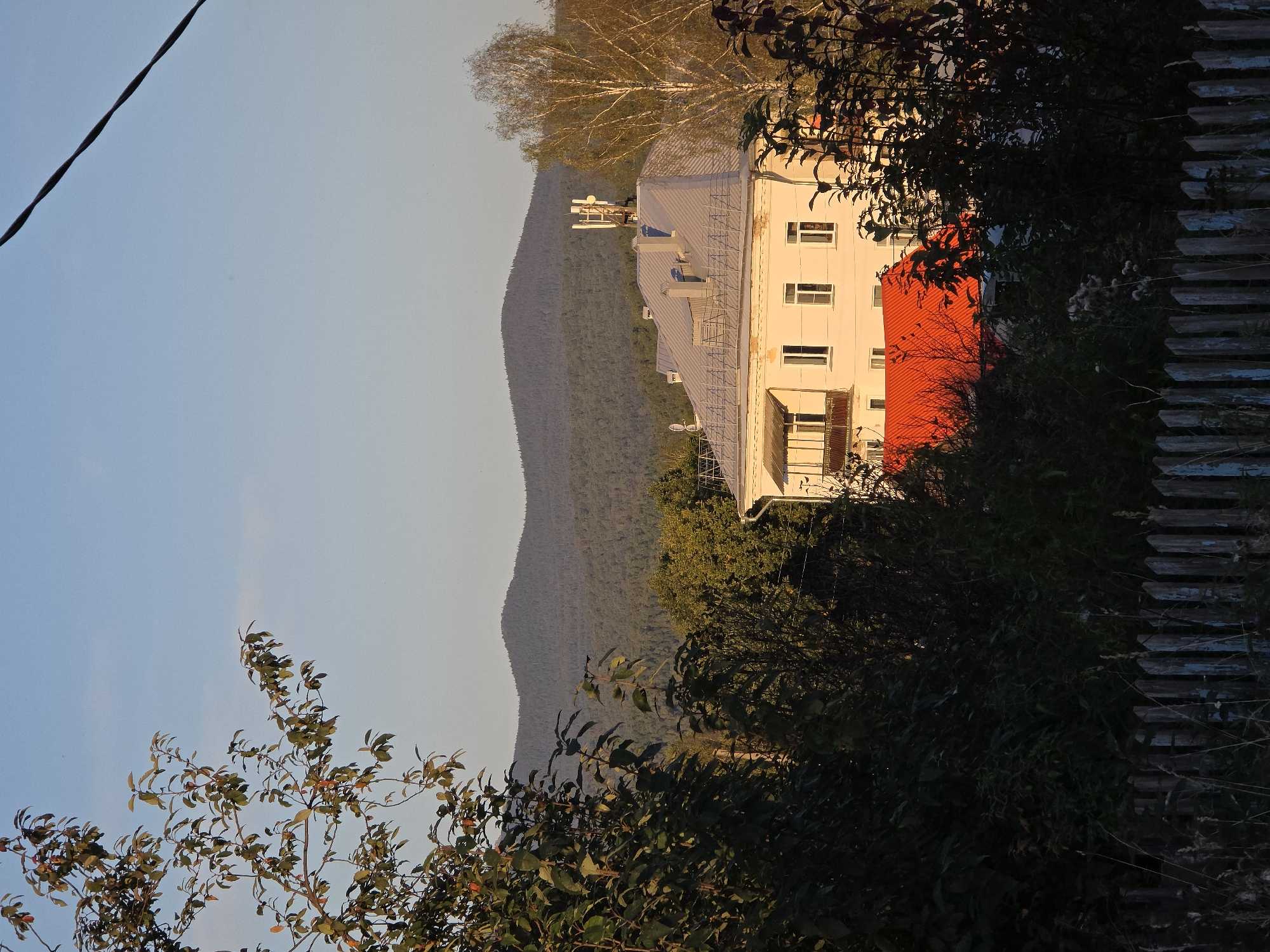 The image depicts a view of a multi-story building with a grey, sloped roof, situated in Magnitka, Russia. The building is light-colored and features multiple windows and a balcony with a railing. A vibrant red-tiled roof is visible on a lower section of the building. On the main roof, there are two chimneys and communication antennas. A tall, thin tree with light-colored bark stands to the right of the building, illuminated by the sun. The background is dominated by rolling, forested hills under a clear blue sky, with a single power line cutting across the upper right quadrant of the image. In the foreground, dense foliage and a weathered wooden fence partially obscure the view. The scene appears to be captured during the late afternoon or early evening, given the warm, golden light. There are no people or animals visible in the image.