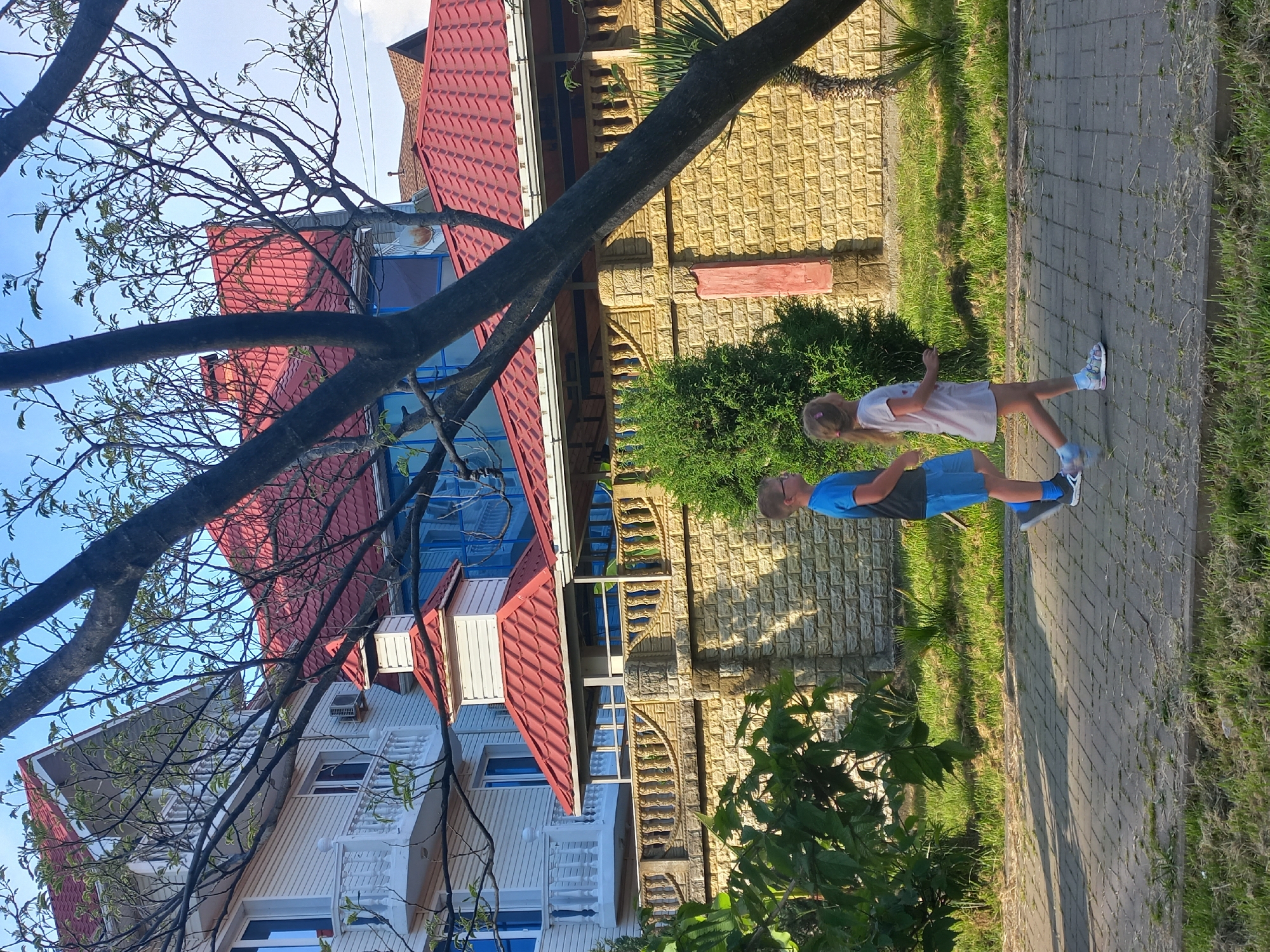 The image shows a residential scene in Unknown, Unknown, featuring a house with a red tiled roof and a beige, textured facade. In the foreground, two children are standing on a paved walkway. The child on the left is wearing a blue shirt and shorts, and the child on the right is wearing a white shirt and shorts. A tree branch obscures a portion of the house, extending from the upper left corner towards the center of the frame. The environment consists of green grass and shrubs along the walkway and in front of the house.