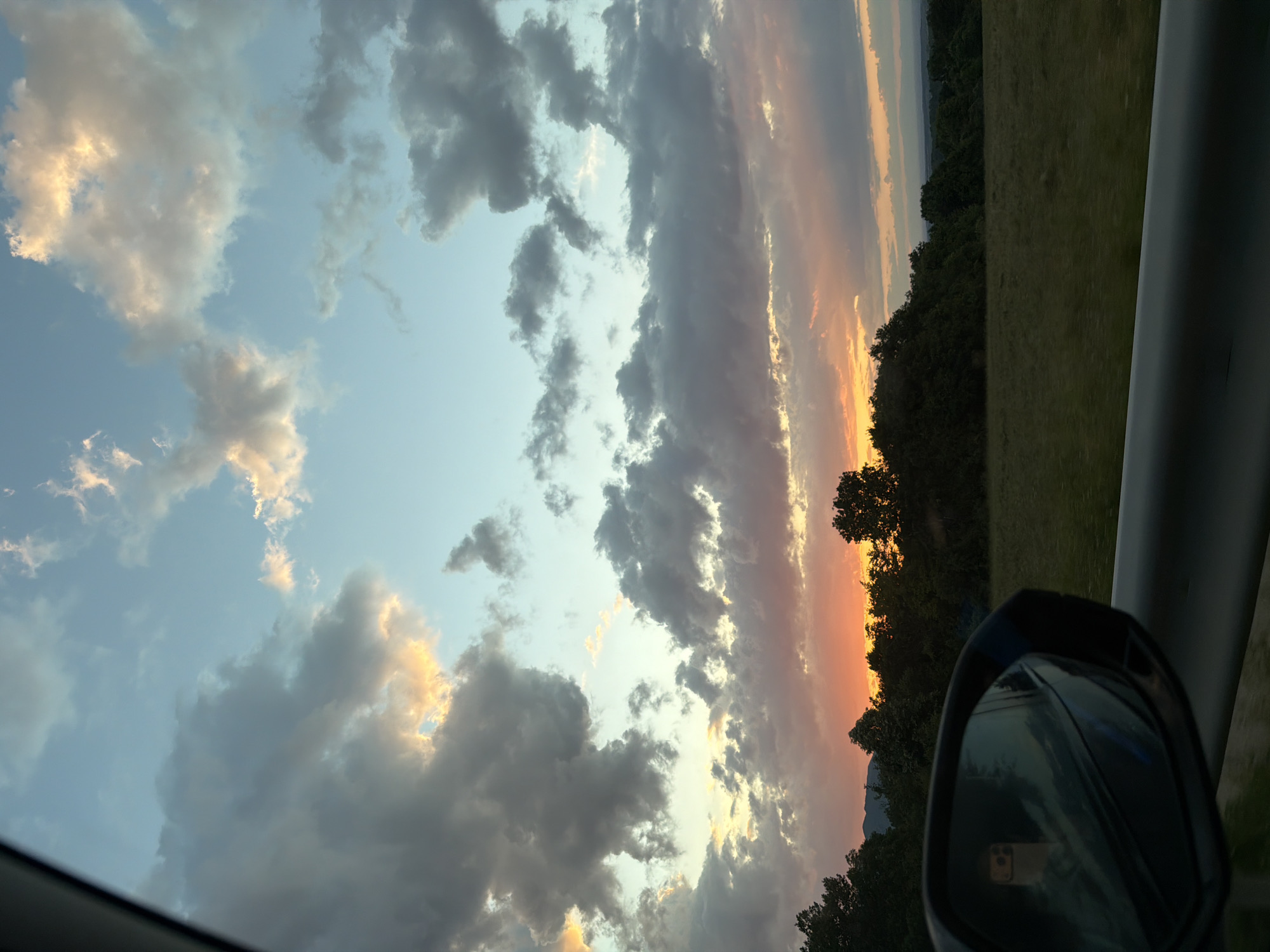 From the interior of a car, the image captures a view of the sky filled with clouds, illuminated by the colors of a sunset. A band of orange and pink light stretches across the horizon, visible above a tree line. To the right of the frame is the side mirror of the car, reflecting part of the scene. The location is Novakovo, Bulgaria.