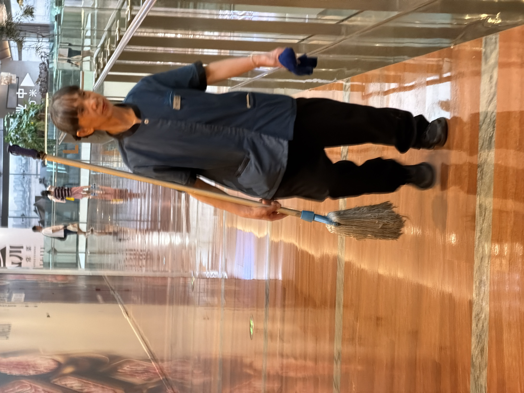 A person is mopping a polished wooden floor in what appears to be a shopping mall or public walkway in Yuzhong District, China. The floor is highly reflective, showing streaks from the mopping action. The person is wearing a dark blue uniform and black pants, and is holding a mop with a white string head and a blue handle. A blue cloth is clutched in their right hand. In the background, other people are visible walking along the walkway, and there are escalators and signage. The environment appears to be indoors, with overhead lighting contributing to the bright reflections on the floor.