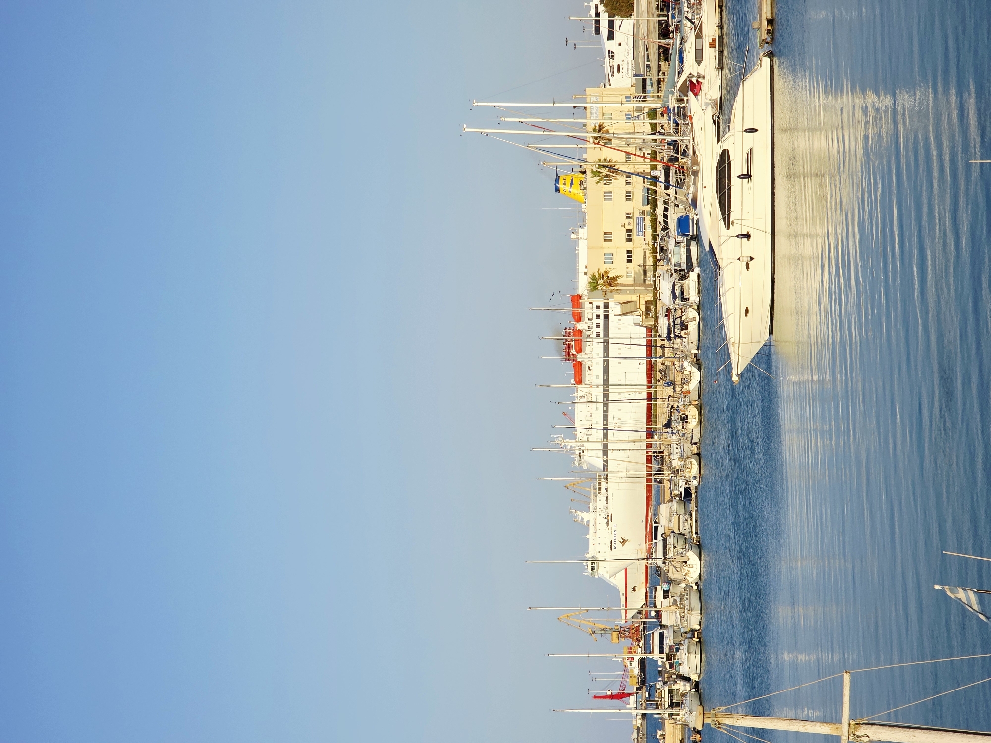 The image depicts a harbor scene in the city of Heraklion Municipal Unit, Greece, under a clear blue sky. In the foreground, calm blue water reflects the scene. Several white sailboats with tall masts are moored, some partially visible. In the mid-ground, a collection of smaller white motorboats and sailboats are docked. Further back, a large white ferry with red trim is prominently docked, identified by the name "ASTERION II" on its side. Adjacent to the ferry are several buildings, one of which is light yellow with numerous windows and a yellow smokestack bearing blue lettering. The background reveals a clear sky and a distant, light-colored structure. The scene is peaceful and suggests a bustling port environment.