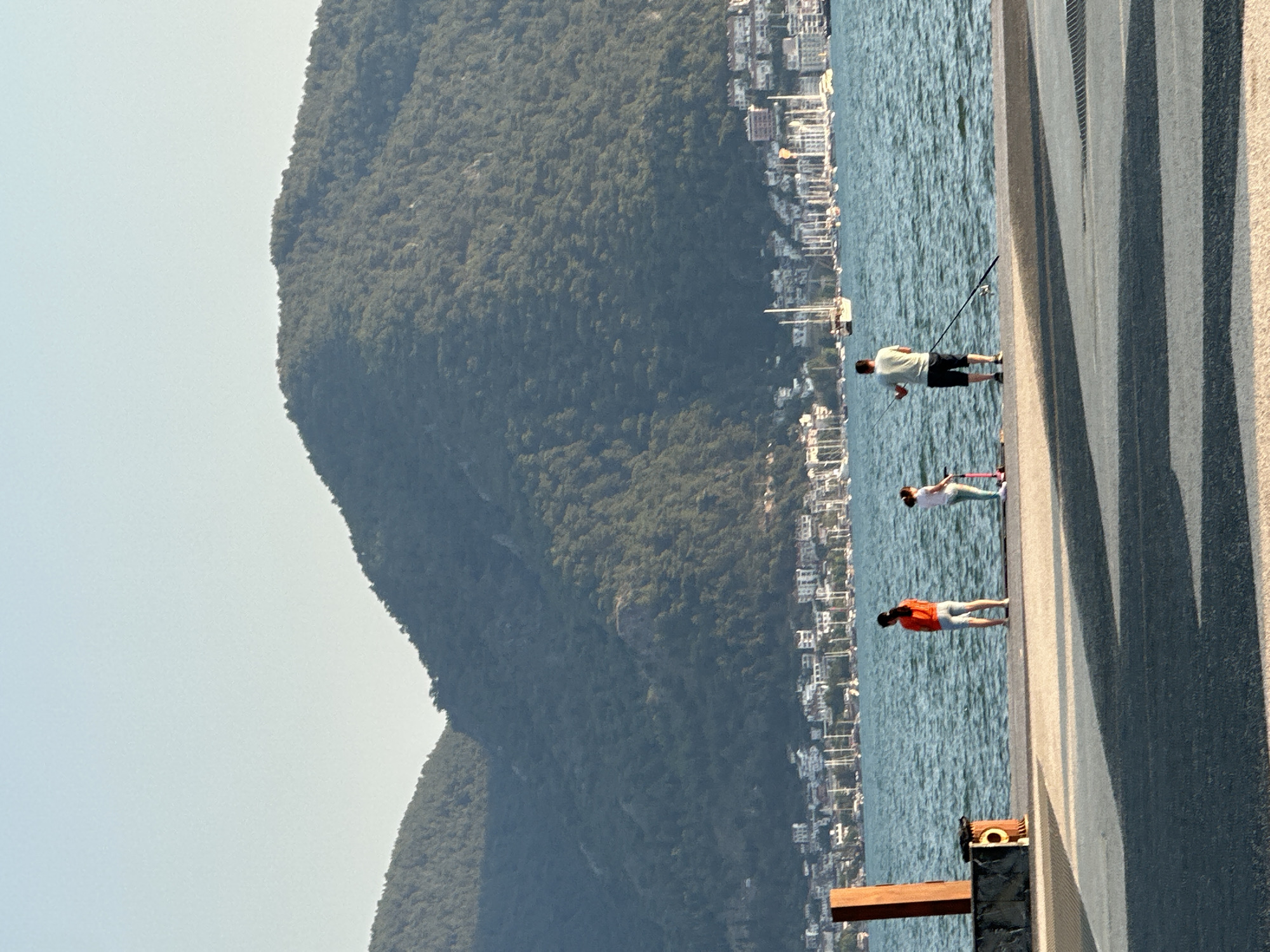 In Fethiye, Turkey, three individuals are positioned on a pier overlooking a body of water. A person wearing an orange shirt and light-colored shorts stands facing the water. To their right, a child on a pink scooter is also looking out at the water. Further to the right, a person in a light-colored shirt and dark shorts is holding a fishing rod, with the line cast into the water. Behind them, numerous sailboats are anchored, and a city with buildings climbs the densely forested slopes of a large mountain. The sky is clear and a pale blue. The pier is paved with a textured surface, and a wooden post stands to the left.
