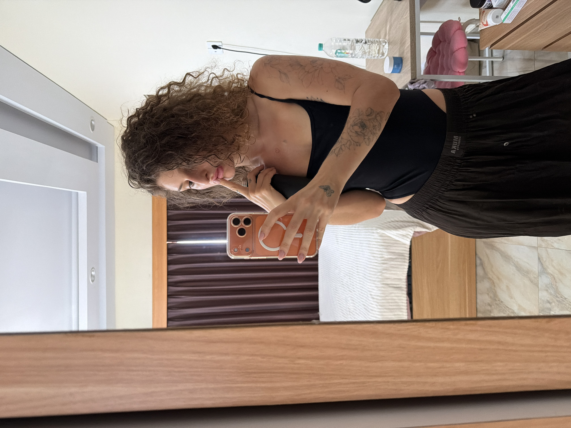 A young woman with curly brown hair is taking a mirror selfie in a room. She is wearing a black tank top and dark brown loose-fitting pants. The room appears to be a bedroom, with a bed visible in the reflection of the mirror. There is a wooden dresser with various items on it, including a bottle of water and some toiletries. The walls are painted a light color, and there are dark brown curtains. The woman is holding an orange phone in her right hand and her left hand is resting on her chin. She has several tattoos on her arms and a small heart tattoo on her chest.
