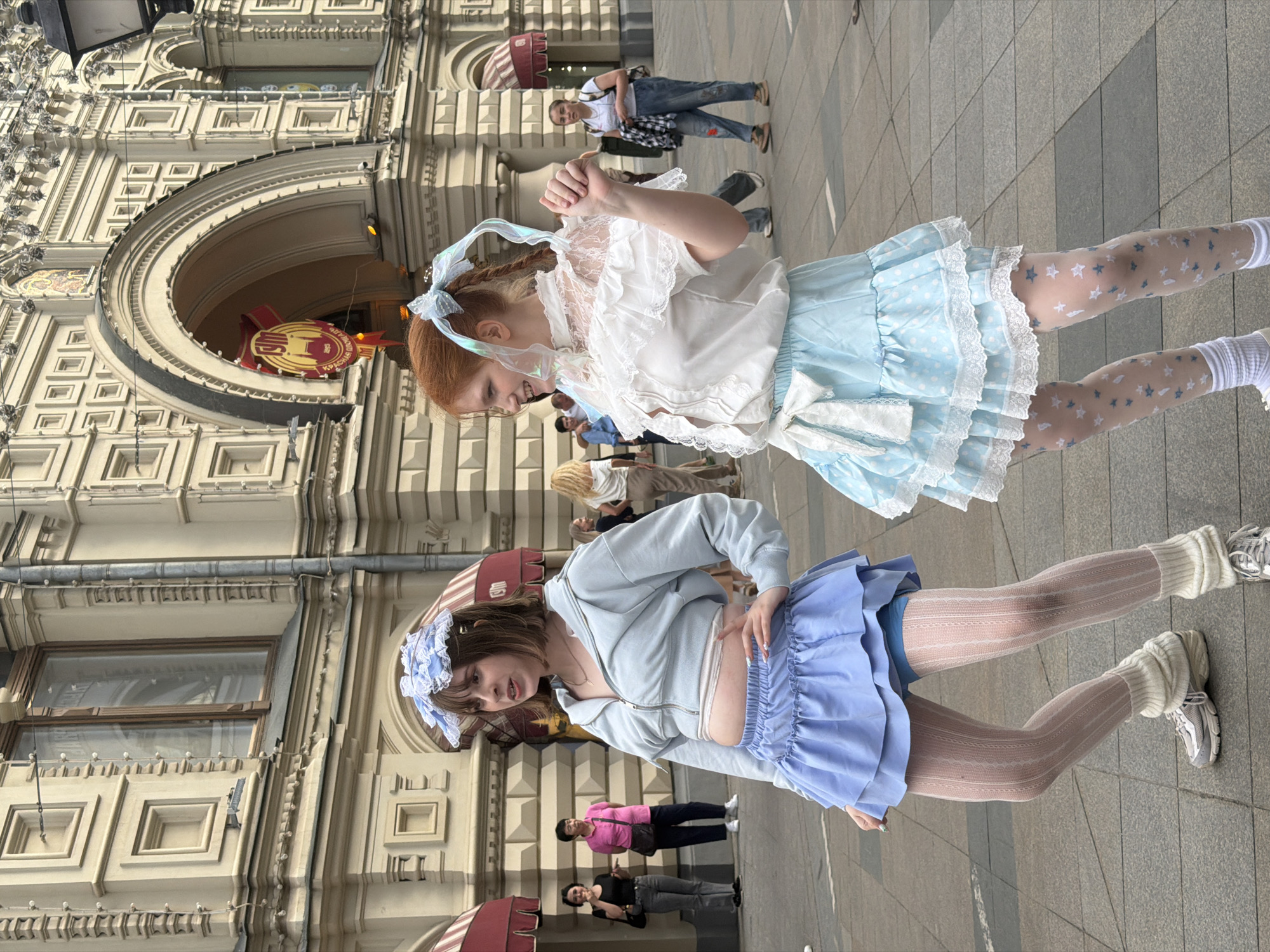 Two young women in complementary blue and white lolita-style dresses pose outdoors. The woman on the right has red hair and a white and light blue dress with star-patterned tights. The woman on the left has brown hair and wears a light blue hooded top with a matching skirt, textured tights, leg warmers, and athletic shoes. They are standing in front of a large, ornate building with arched windows, identified by a Cyrillic sign. The background includes other pedestrians and architectural details typical of Moscow, Russia.