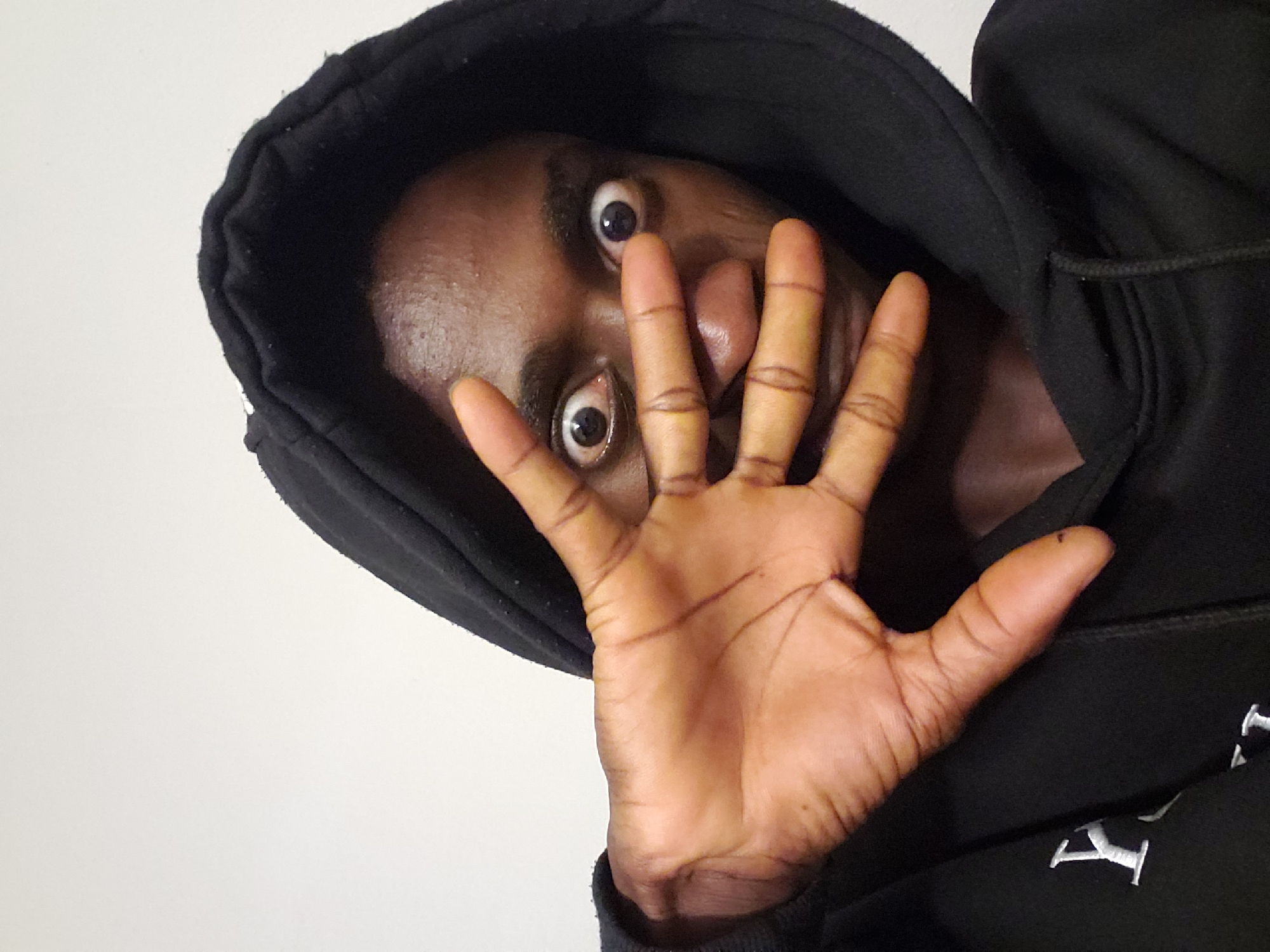 A person with dark skin, wearing a black hooded sweatshirt, is pictured looking downward toward the camera with wide eyes. The hood is pulled up, framing the face. The person's right hand is held up, partially covering the face. The background is a plain light color, possibly a wall. The photograph is taken from a low angle. There is no indication in the image that it was taken in the city of Adonkolo, Nigeria.