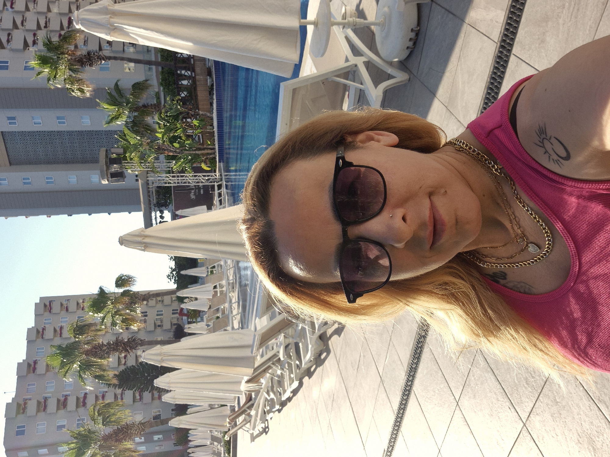 The image shows a woman taking a selfie outdoors at a resort. The woman has blonde hair, is wearing sunglasses, a pink tank top, and gold necklaces. She has a tattoo on her right shoulder. In the background, there is a swimming pool, lounge chairs, and umbrellas. Behind the pool area, there is a large, multi-story building with balconies, and palm trees. The scene appears to be a sunny day.