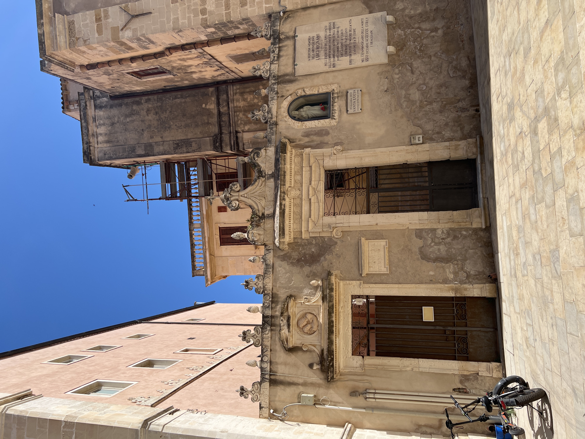 The image shows a street scene in Syracuse, Italy, likely in the historic center. In the foreground, two electric scooters are parked on a paved courtyard made of light-colored rectangular stones. The scooters are dark-colored with some blue accents.
The background features an old building with a beige facade. The building has two distinct entrances, both with ornate architectural details and heavy metal gates. The left entrance has a framed circular relief above it, while the right entrance features a small niche with a statue of a Madonna and child, illuminated by a red flower placed at its base. Above the right entrance, a plaque is mounted on the wall with text in Italian. Another, smaller plaque is visible near the right entrance. The facade of the building exhibits signs of age and weathering, with some discoloration and patches of wear.
To the left of this building, a taller, modern structure with a pinkish-orange facade is visible, with several rectangular windows. To the right, another older building with stone construction is partially visible, with a terracotta-tiled roofline and scaffolding attached to one section, suggesting ongoing restoration work. The sky above is a clear, deep blue. The overall impression is one of historical architecture bathed in sunlight, typical of a Mediterranean city.