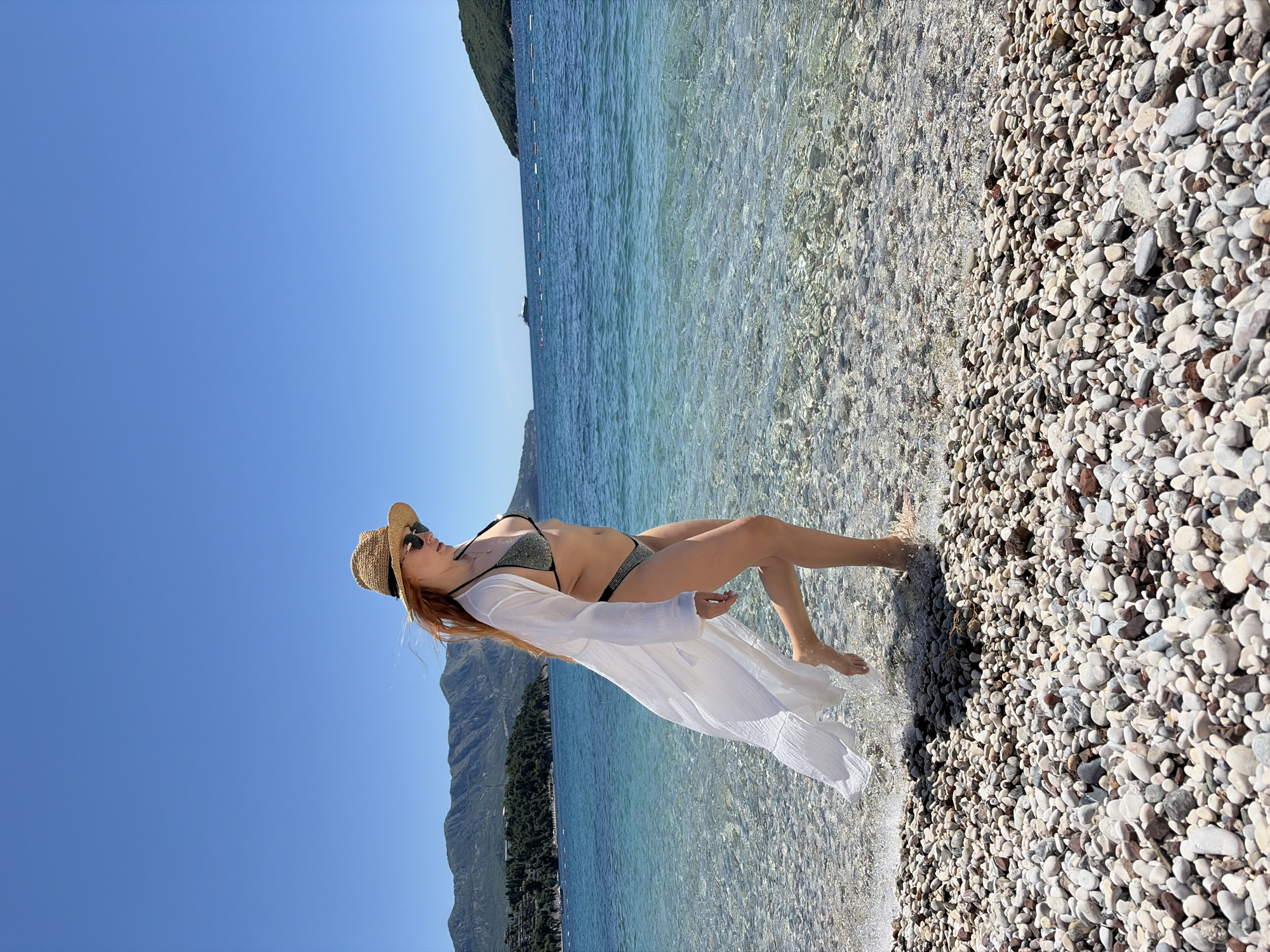 The image depicts a woman standing on a pebble beach at the edge of the water in Budva, Montenegro. She is wearing a bikini, a straw hat, sunglasses, and a long white cover-up. Behind her is the turquoise-colored water of the Adriatic Sea and a backdrop of mountains. The sky is clear and blue. The beach is made up of small, smooth pebbles of various colors.