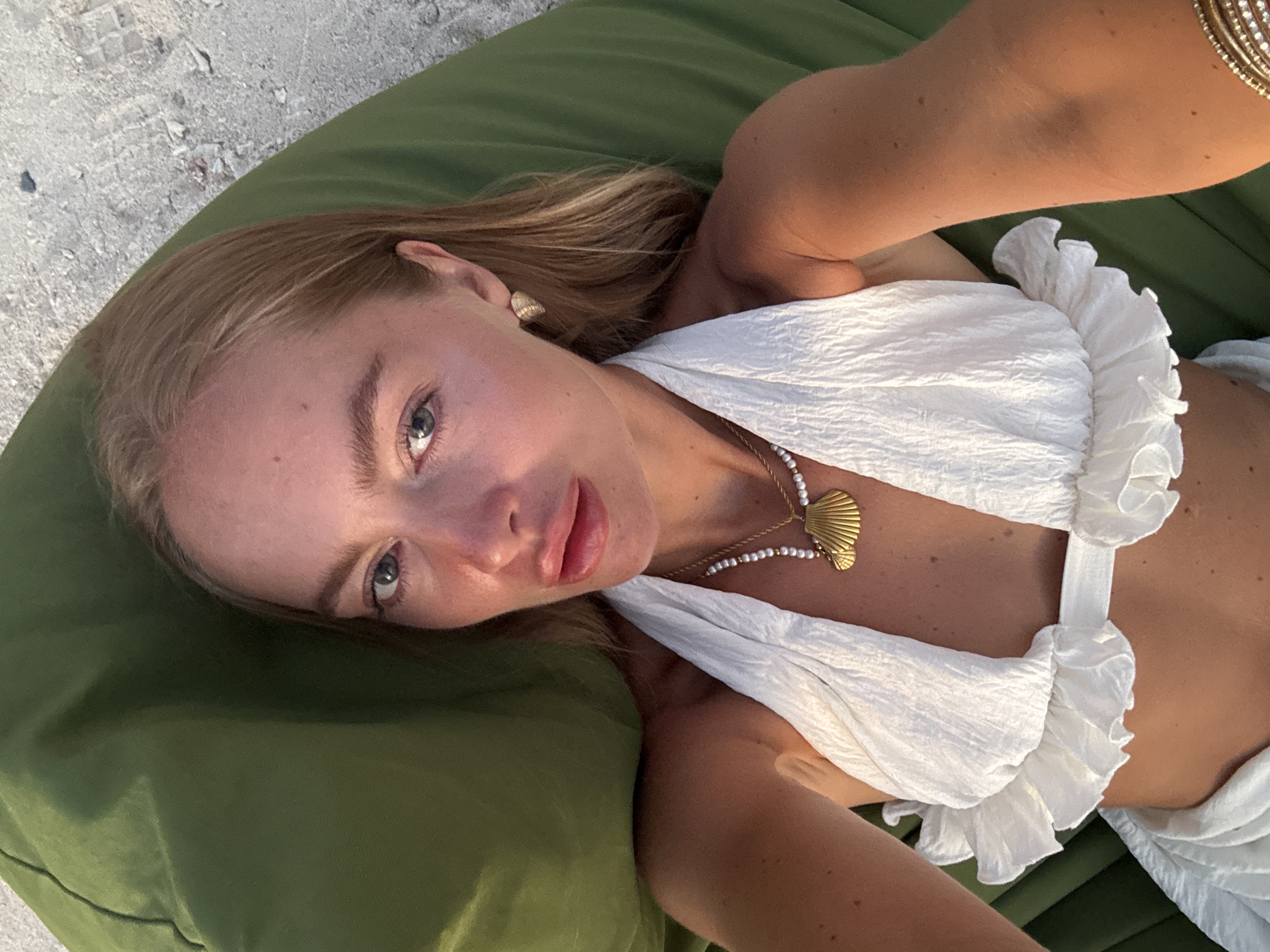 A blonde-haired woman with fair skin is laying on an olive-green cushion or beanbag. She is wearing a white halter top with ruffles. A gold necklace with a seashell pendant is visible on her chest. She also has gold earrings and a bracelet. The background shows a sandy surface, suggesting a beach setting. The location is Unknown, Indonesia.
