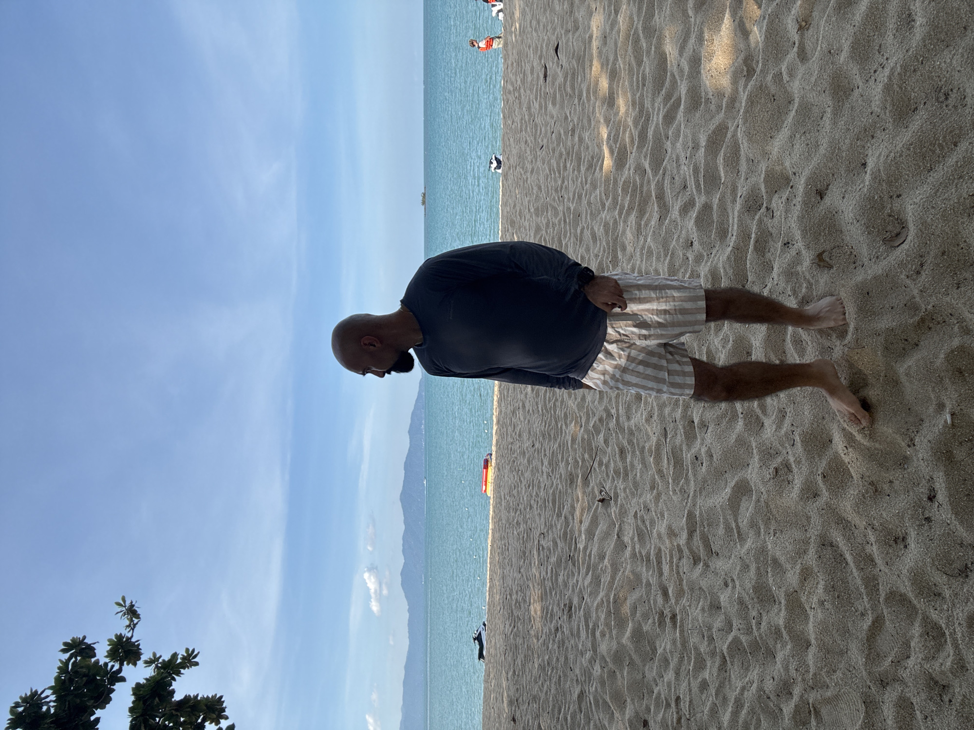 An adult bald man with a beard stands on a sandy beach in Ko Samui District, Thailand, looking towards the ocean. He is wearing a long-sleeved dark blue shirt and striped shorts. The sand is textured with ripples and a few small leaves. The ocean is a vibrant turquoise, with calm waves. In the distance, a line of mountains forms the horizon, and a few boats are visible on the water. The sky is clear blue with a few scattered white clouds. To the left of the frame, a tree branch with green leaves hangs over the scene, casting a shadow on the sand. The overall atmosphere is serene and relaxed, suggesting a pleasant day at the beach.