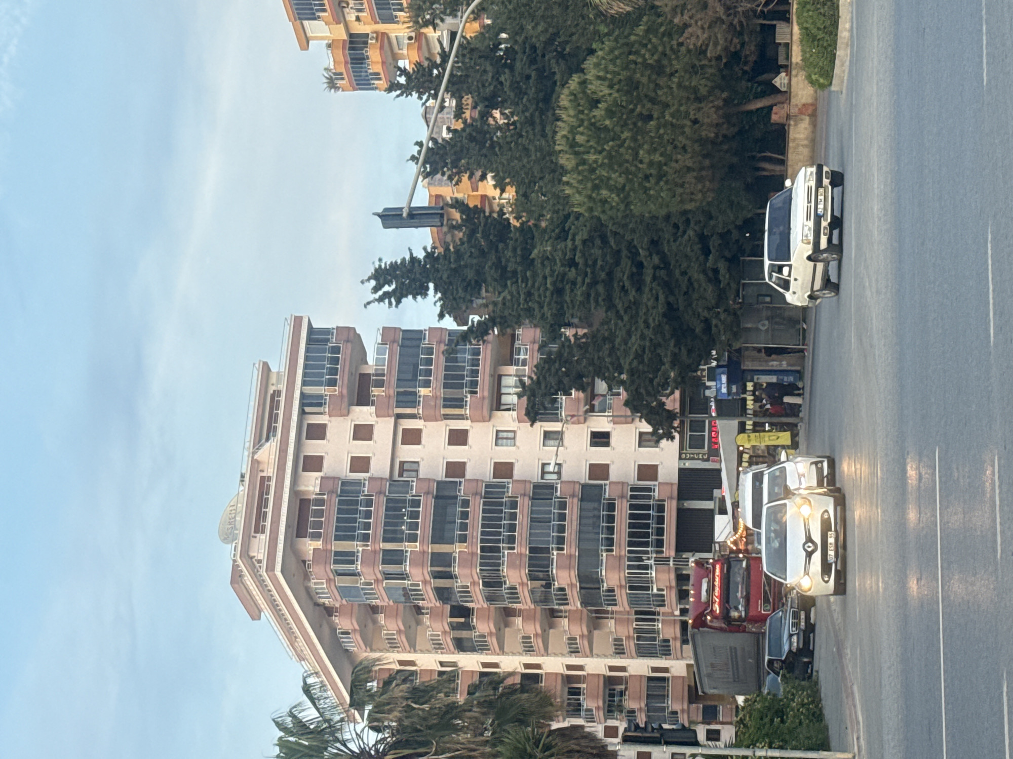 The image captures a street scene in the city of Unknown, Unknown, with a tall, multi-story building prominently displayed on the left. The building features a light beige facade with rows of windows framed in brown. A sign with the name 'KASFET' is visible on the top.
The right side of the image is dominated by a road with multiple vehicles. These include a white car with its headlights on, a red truck, and a white sedan. In the background, there are trees and another building with a similar architectural style but colored in orange. The sky is partially visible and light blue.