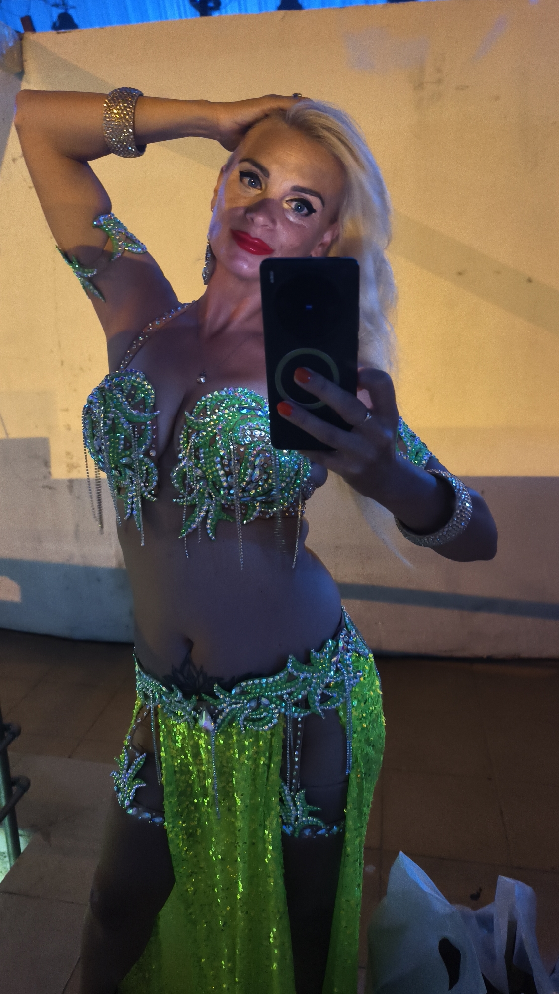 In Kuşadası, Turkey, a woman with blonde hair and bright red lipstick poses for a selfie in a sparkling green belly dance costume. She is wearing a bra-like top adorned with green sequins and clear rhinestones, along with a matching belt and a flowing skirt that features dramatic side cutouts. Her arms are decorated with glittering bracelets, and she has a large, jeweled cuff on her right wrist. She holds a black smartphone in her right hand, capturing her reflection. The background appears to be an outdoor setting with a textured wall and a hint of blue light from above.
