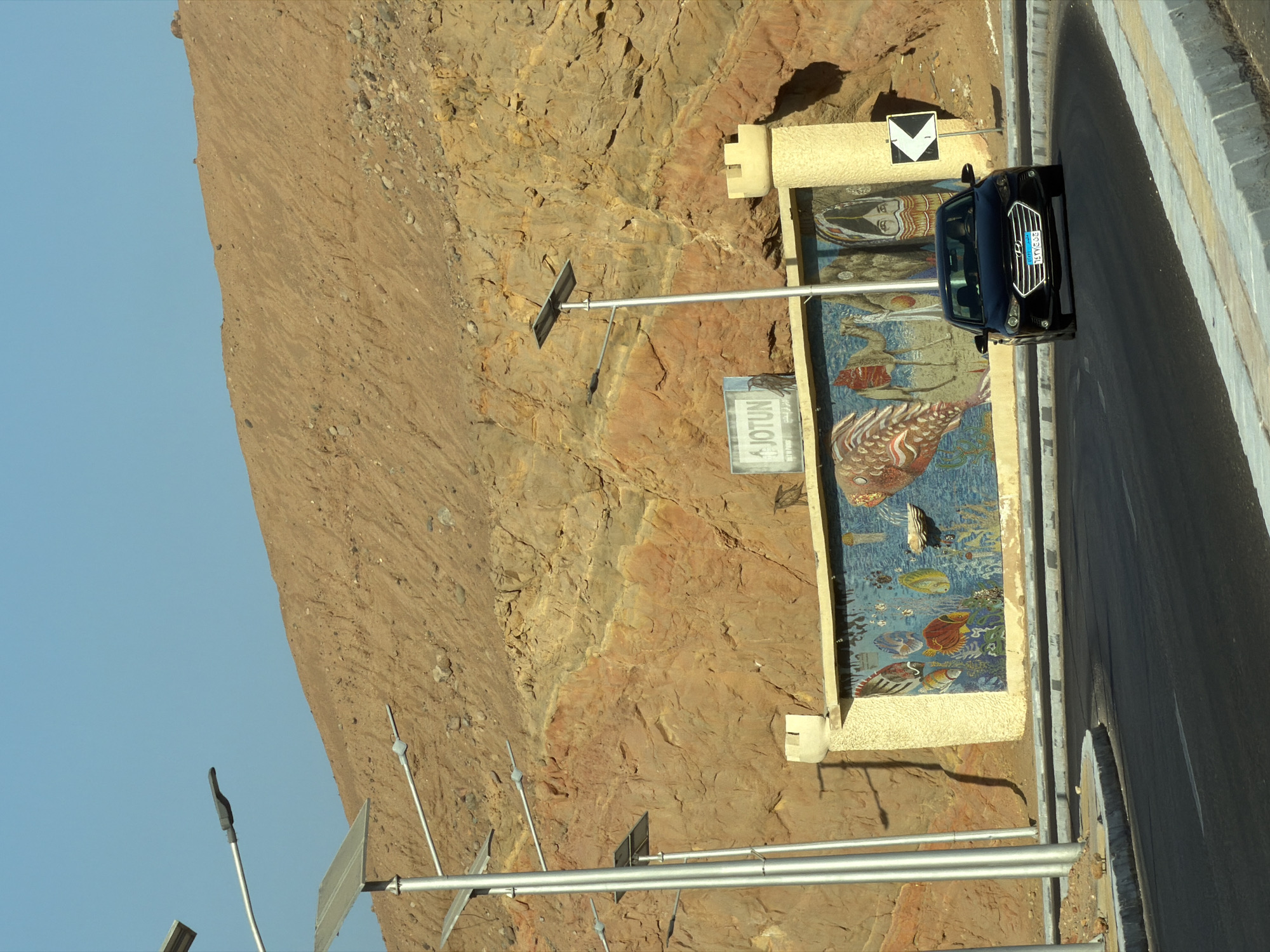The image shows a black Hyundai Elantra driving on a road in Sharm El Sheikh, Egypt. The car is turning to the right. The road is asphalt with a white line dividing the lanes and a concrete curb. Behind the road is a wall with a mosaic mural depicting fish, a camel, and a woman in traditional attire. Above the wall is a rocky hill. There are solar-powered streetlights along the road. The sky is clear and blue.