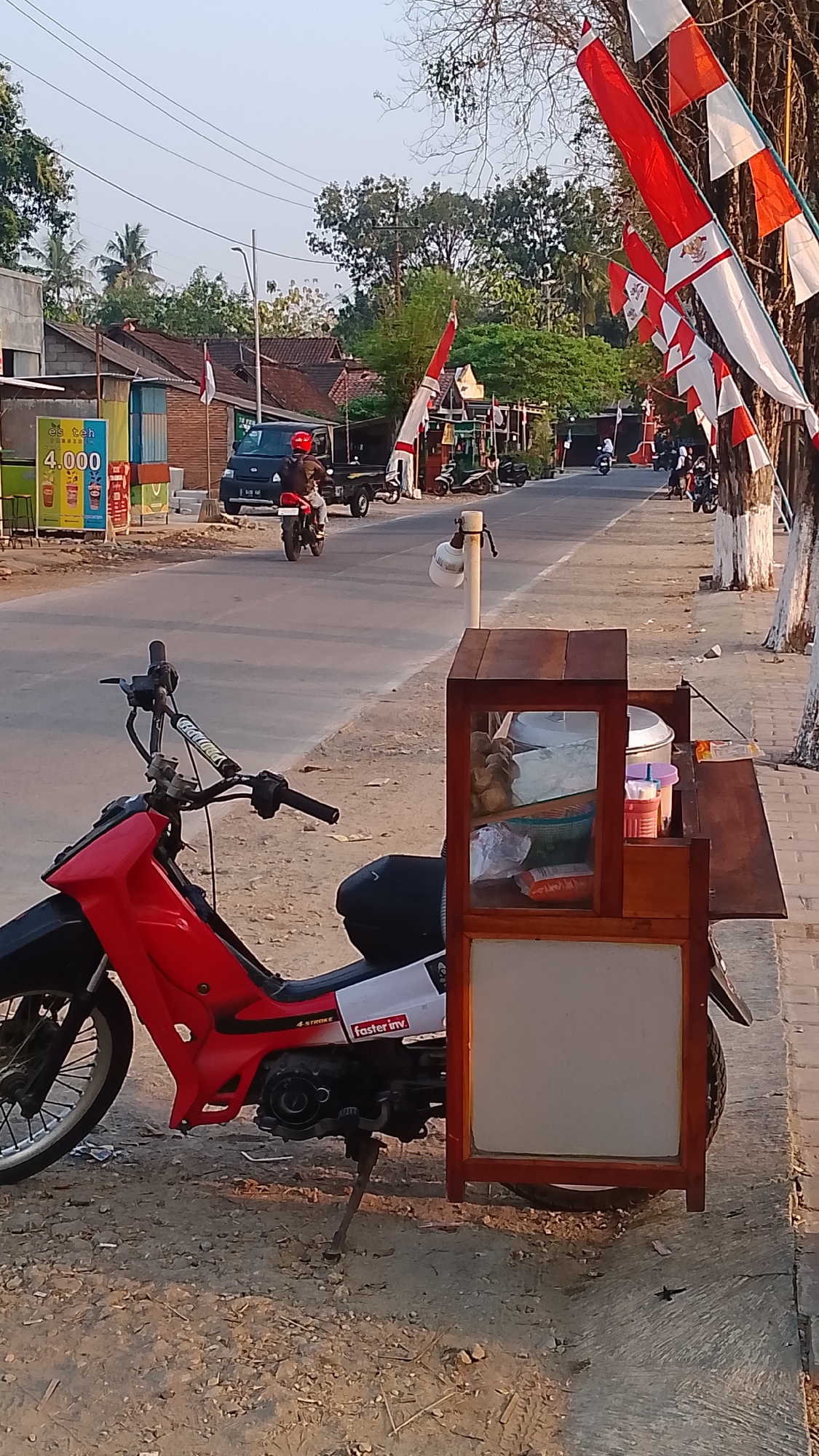 The image depicts a street scene in Karanglangu, Indonesia, likely during the late afternoon or early evening due to the warm, low-angle sunlight. In the foreground, a red and black motorcycle is parked, equipped with a wooden cart attached behind it. The cart appears to be a mobile food or drink stand, with glass panes and shelving displaying various items, including containers and what seem to be snacks. The motorcycle has "4 STROKE" and "faster inv." decals.
The street itself is paved, with a gravel shoulder on the right side where the motorcycle and cart are parked. Along the right side of the street, there are several trees with white-painted bases, and decorative Indonesian flags (red and white) are strung between them and other poles, suggesting a festive or patriotic occasion.
In the background, a black pick-up truck is visible on the road, with a rider on a motorcycle behind it. Further down the street, more motorcycles are parked, and several people are seen walking or riding. Buildings line the left side of the street, some with visible awnings and signs, including one with "4.000" printed prominently. The overall environment suggests a moderately busy roadside location within a town or village.