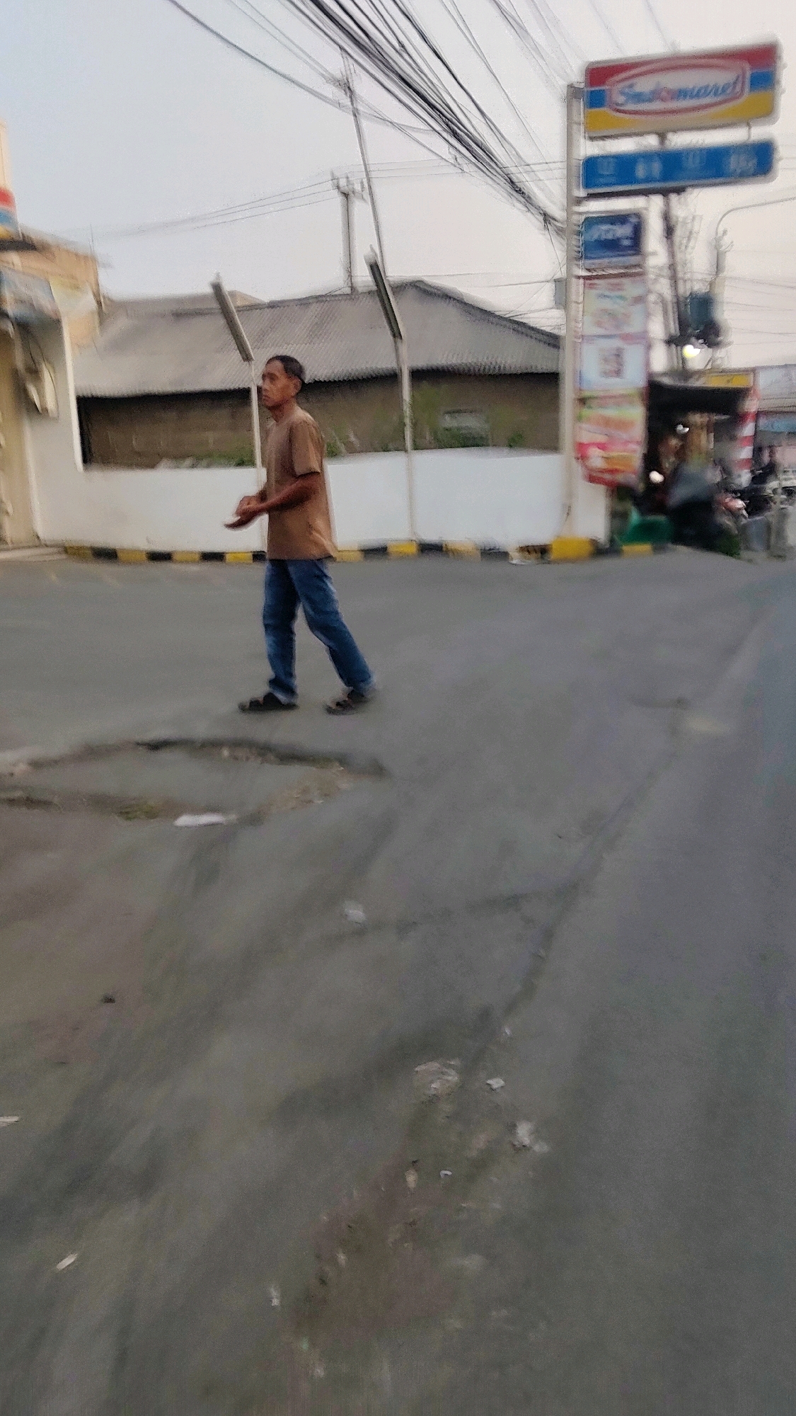 A man walks on a paved road in front of a building with a grey corrugated roof. He wears a tan t-shirt and blue jeans with black sandals. He is holding a small, round object in his hands. To the right of the man is a signpost with the logo "Indomaret" and other smaller signs. The road has several potholes. The sky is overcast.