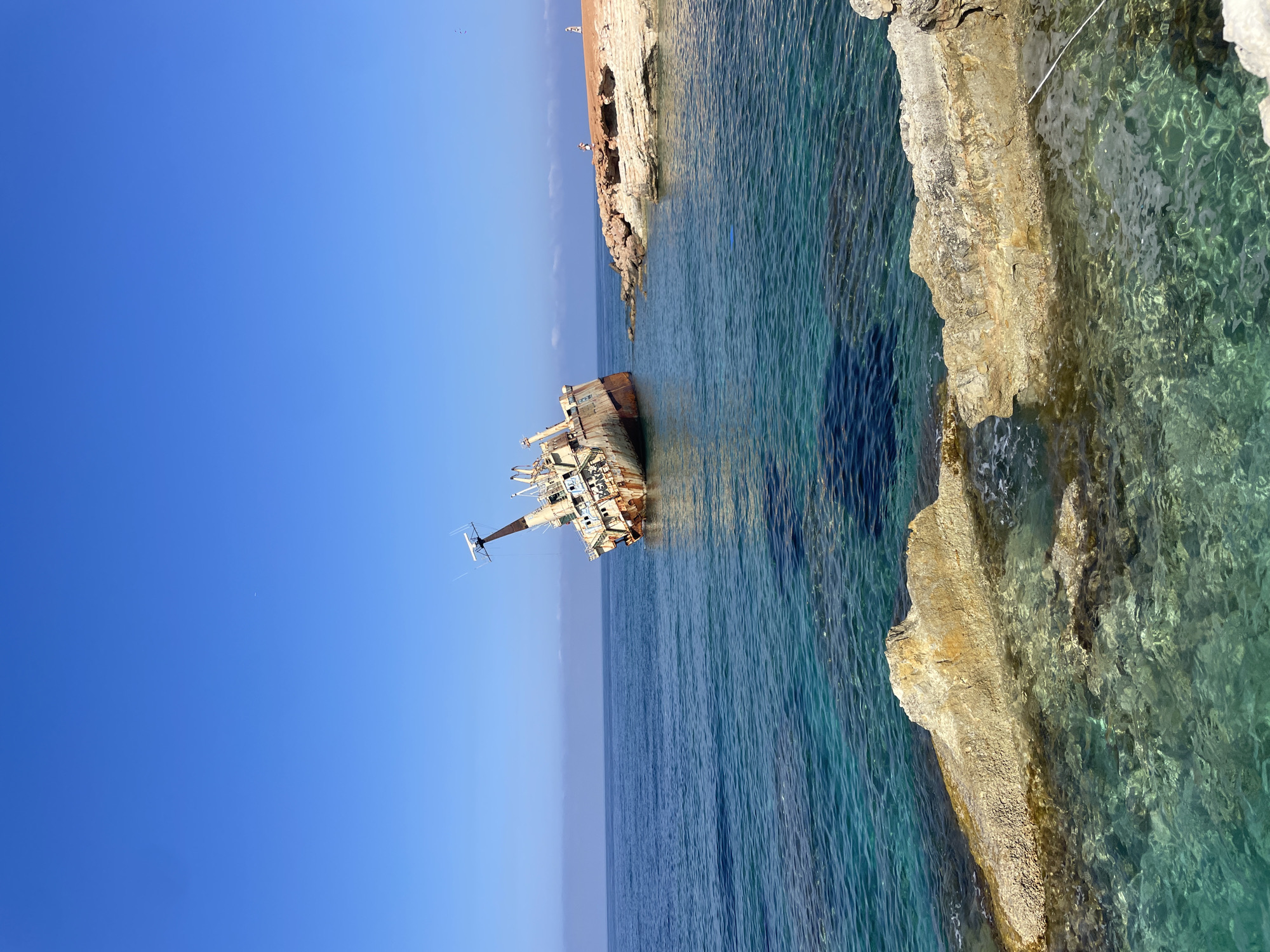 This is a photograph of the shipwreck of the EDRO III, located in the sea near Peyia, Cyprus. The rusty, beached cargo ship lists dramatically to its port side. The ship is partially submerged in clear, turquoise water, with its stern resting on the seabed and its bow pointing upwards. In the background, the sky is a vibrant blue with a few distant white clouds. The foreground shows a rocky shoreline with the shallow, clear water revealing the seabed below. On the rocky outcrop to the right of the shipwreck, two small figures can be seen standing, appearing as tiny specks against the vastness of the sea and sky. The overall scene depicts a dramatic maritime disaster resting in a picturesque Mediterranean setting.