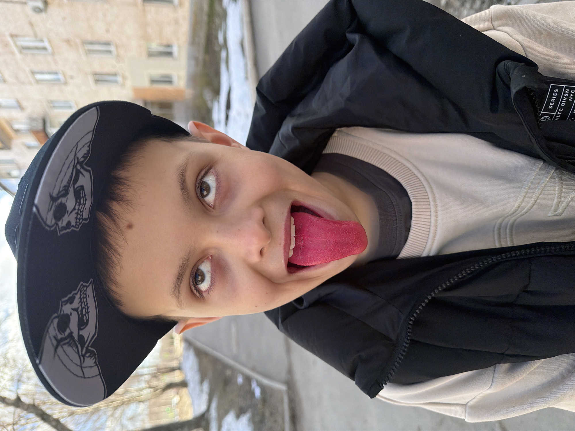 A photograph, taken outdoors, depicts a boy. The boy wears a baseball cap with a skull design, a light-colored crew neck sweater, and a black jacket. He is sticking his tongue out. The background shows an urban environment, possibly Votkinsk, Russia, with a building and patches of snow on the ground.