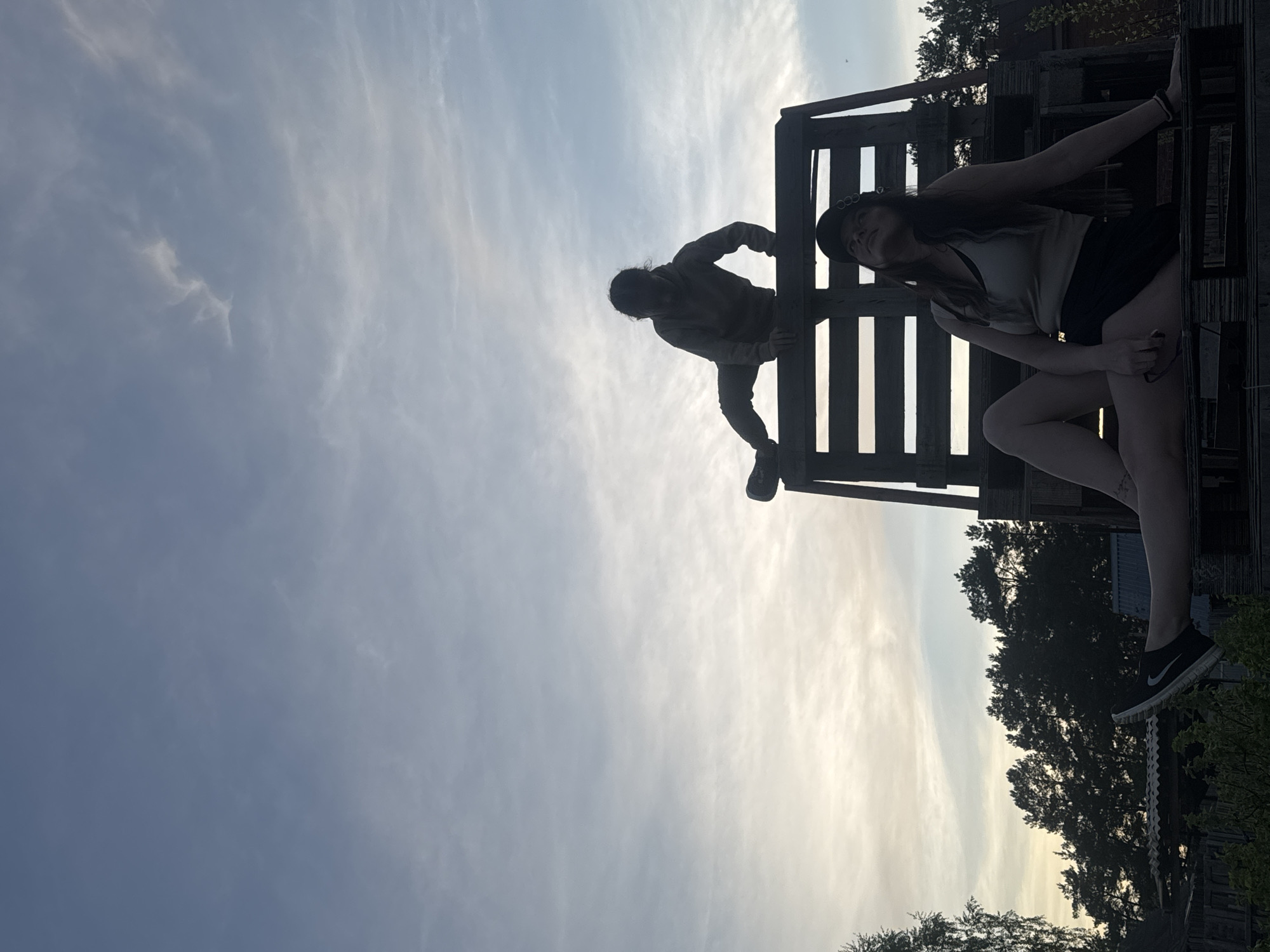 Two people are positioned on a wooden structure outdoors. One person is climbing on the structure while the other person is sitting atop it. The scene is set against a sky with clouds and trees in the background. The location is Rusalevka, Russia.
