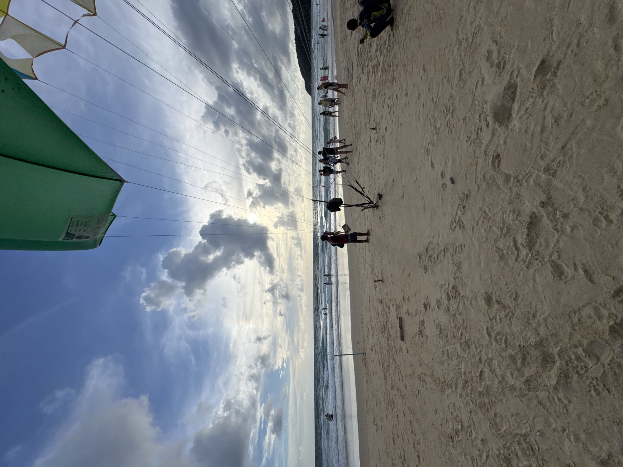 The image depicts a beach scene at Karon, Thailand. In the foreground, a sandy beach stretches towards the ocean, with footprints visible in the sand. Several people are present on the beach. In the midground, the ocean has moderate waves and a few individuals are in the water. The sky is partly cloudy with the sun peeking through, creating a bright, diffused light. A large green parachute is visible in the upper portion of the image, with numerous ropes extending downwards, suggesting a paragliding activity is taking place or about to commence. Several individuals appear to be involved in setting up or preparing for this activity. The overall atmosphere is one of a recreational beach setting on a day with dynamic weather conditions.