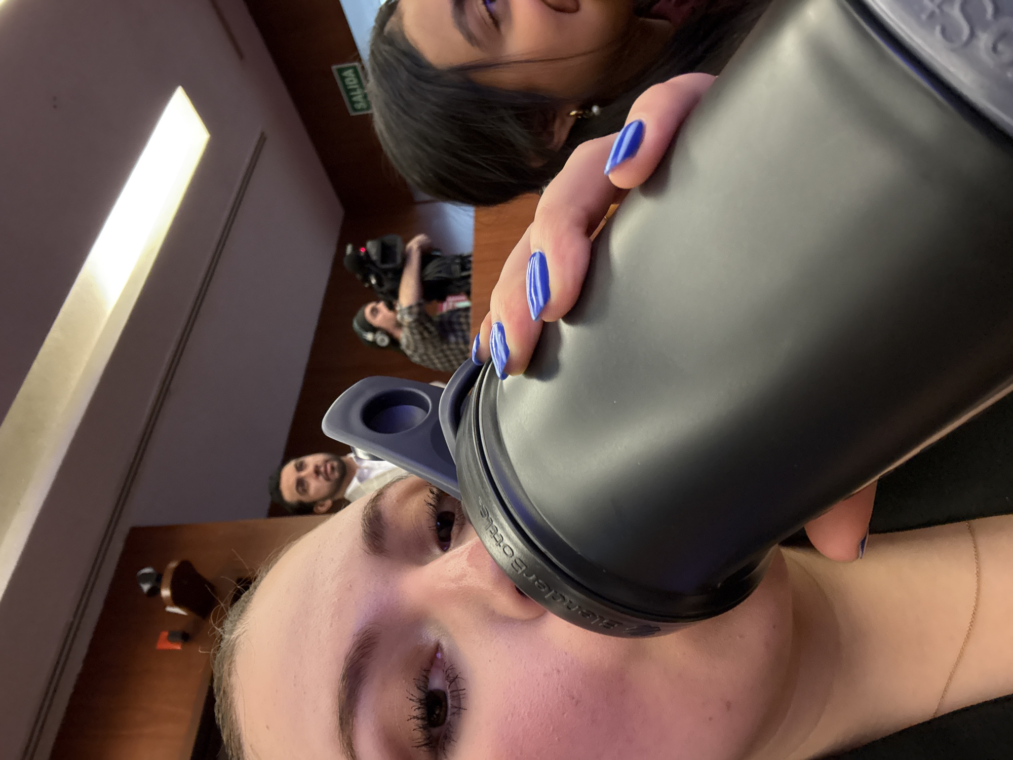 An indoor scene, likely a public or event space in Mexico City, Mexico, is captured from a low-angle, first-person perspective. In the immediate foreground, a female subject's lower face and right hand are visible as she drinks from a dark gray BlenderBottle. Her right hand displays blue-painted fingernails. A thin gold chain is present around her neck. In the background, multiple individuals and environmental details are discernable. To the left, a male subject with dark hair and a beard, dressed in a light-colored suit jacket over a white shirt, observes an unseen focal point. Behind him, another male with long dark hair, wearing a patterned shirt and headphones, operates a black professional video camera. To the right, part of a female subject's face is visible, showing dark hair, a pearl earring, and a slight smile. The environment includes wooden wall paneling, a rectangular ceiling light fixture, and a green "SALIDA" (EXIT) sign with white text. A small, dark, wall-mounted device is positioned on a wooden bracket on the left wall.