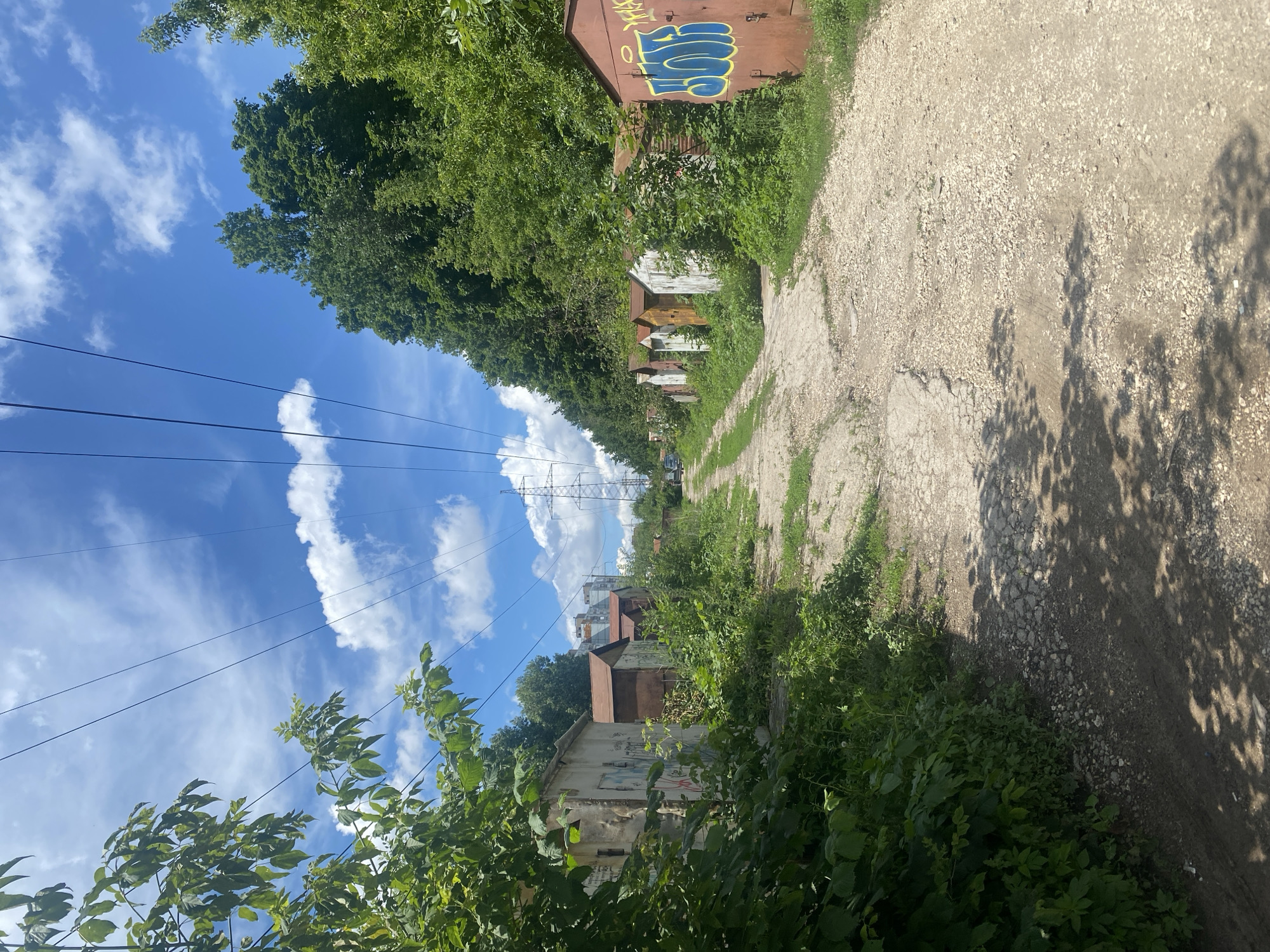 This is a street-level, vertical, eye-level shot of a dirt road lined with garages on both sides. The road appears to be a path leading away from the viewer, with a power line tower visible in the distance. The garages are old and appear to be in disrepair, with some showing signs of graffiti. The road is cracked and covered with gravel, and there is vegetation growing on either side of the road and between the garages. The sky is blue with scattered white clouds. The scene is a residential area, likely in a developing part of the city of Samara, Russia, characterized by a mix of old garages and newer buildings in the background. There are no people or vehicles visible in the immediate foreground, but a car can be seen in the distance. The lighting suggests it is daytime.