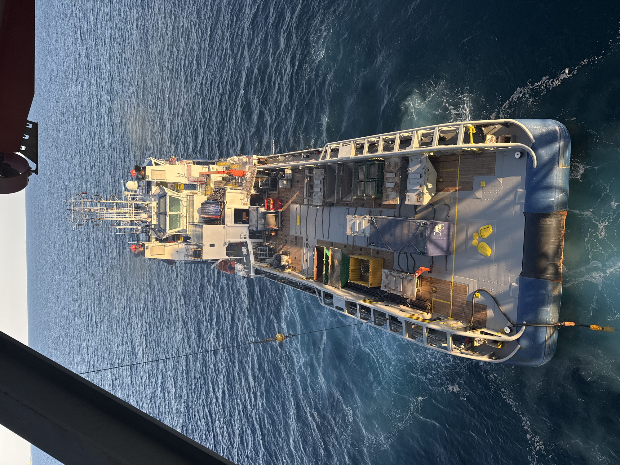An overhead view shows a supply vessel named "IEVOLI BLUE" at sea. The vessel is long and narrow, with a large deck area filled with various equipment and cargo containers. A crane extends from the left side of the image, with a yellow ball suspended from it. The sea is a deep blue with whitecaps. The sky is a light blue with a hint of haze. The vessel appears to be at anchor or moving slowly. The overall scene suggests a maritime operation in progress. There are no buildings or city elements visible, indicating the location is at sea, not in a city in Italy or elsewhere.