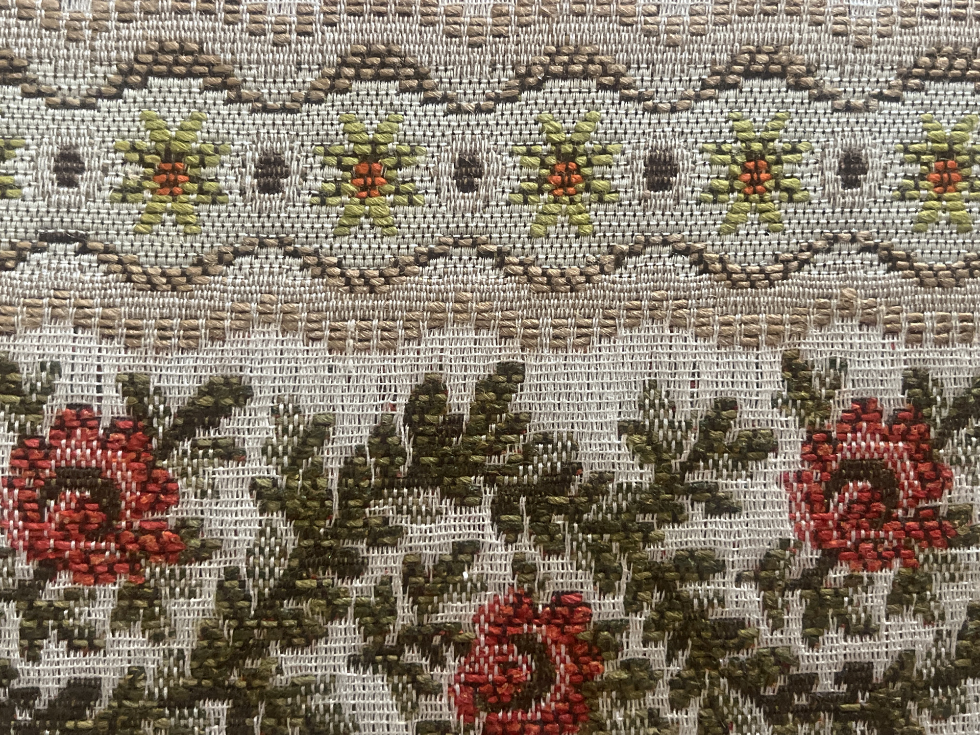 This image presents a detailed close-up of a woven textile, displaying two distinct patterned sections. The left portion features a repeating floral pattern against a light grey or off-white background weave. This pattern consists of stylized red roses, ranging in hue from deep red to an orange-red, accompanied by textured dark green leaves and stems. The weaving technique renders a pixelated or cross-stitch-like appearance for these motifs. The right portion of the textile exhibits a vertical decorative border. This border is composed of a central column, also on a light background, containing repeating small, stylized floral designs with light green petals and orange centers, interspersed with dark brown rectangular elements. This central column is flanked by vertical strips of a coarse, plain beige-brown weave, which are further bordered by a zigzag pattern in a darker brown. The entire textile shows visible individual threads, indicating a coarse-to-medium weave. No people, actions, or specific environmental details are discernible. The image provides no visual information to determine its geographical origin or location within the city of Casablanca, Morocco.