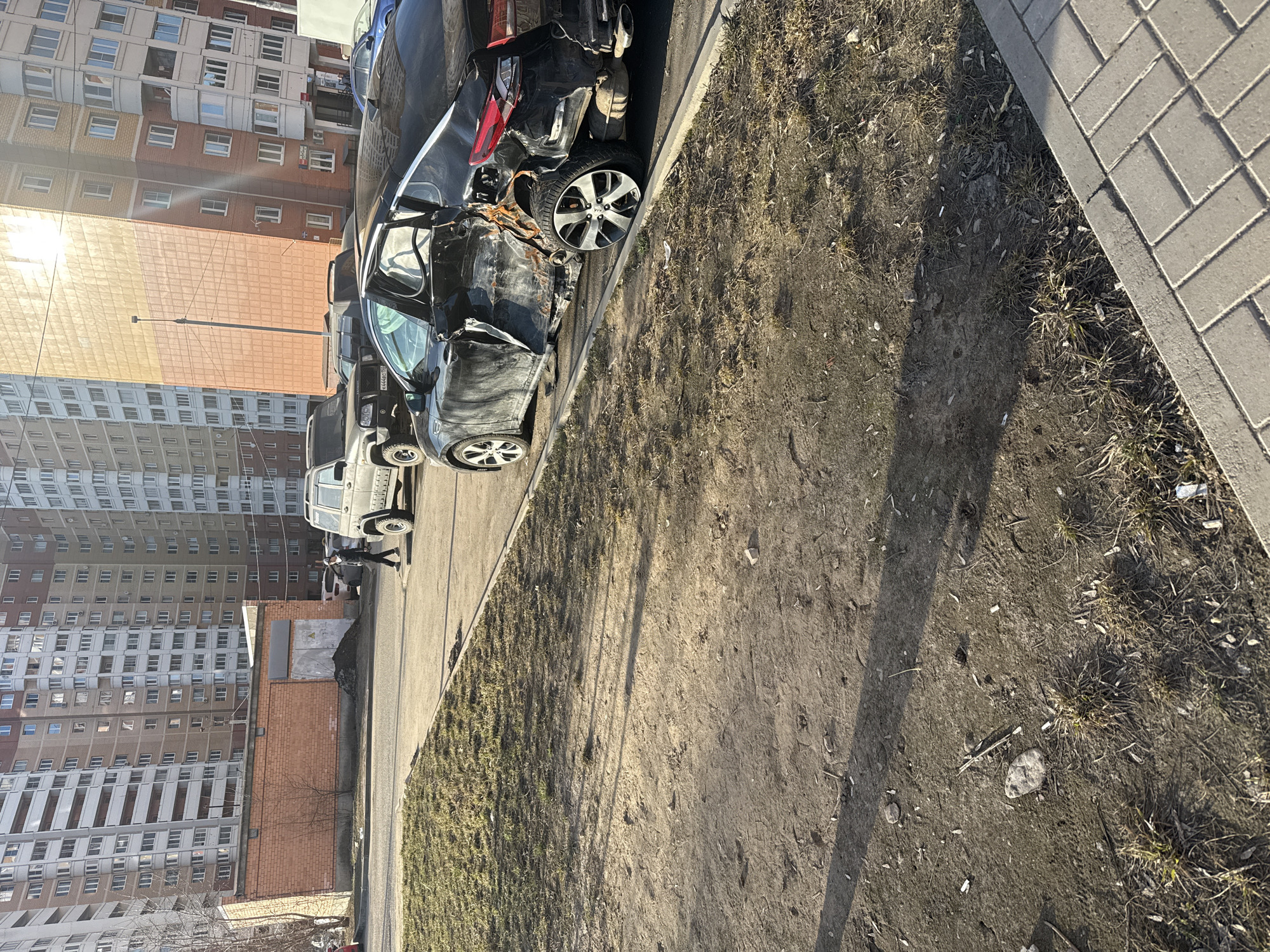 The image depicts a street scene in Dmitrov, Russia, showing a heavily damaged black car parked along the side of a road next to a sidewalk. The front driver's side of the car is severely crushed. Several other vehicles are parked further down the road. A tall apartment building is visible in the background. The ground area between the road and sidewalk is a mix of dirt, sparse grass, and some debris. A lamppost stands nearby. A person in dark clothes can be seen walking in the distance.