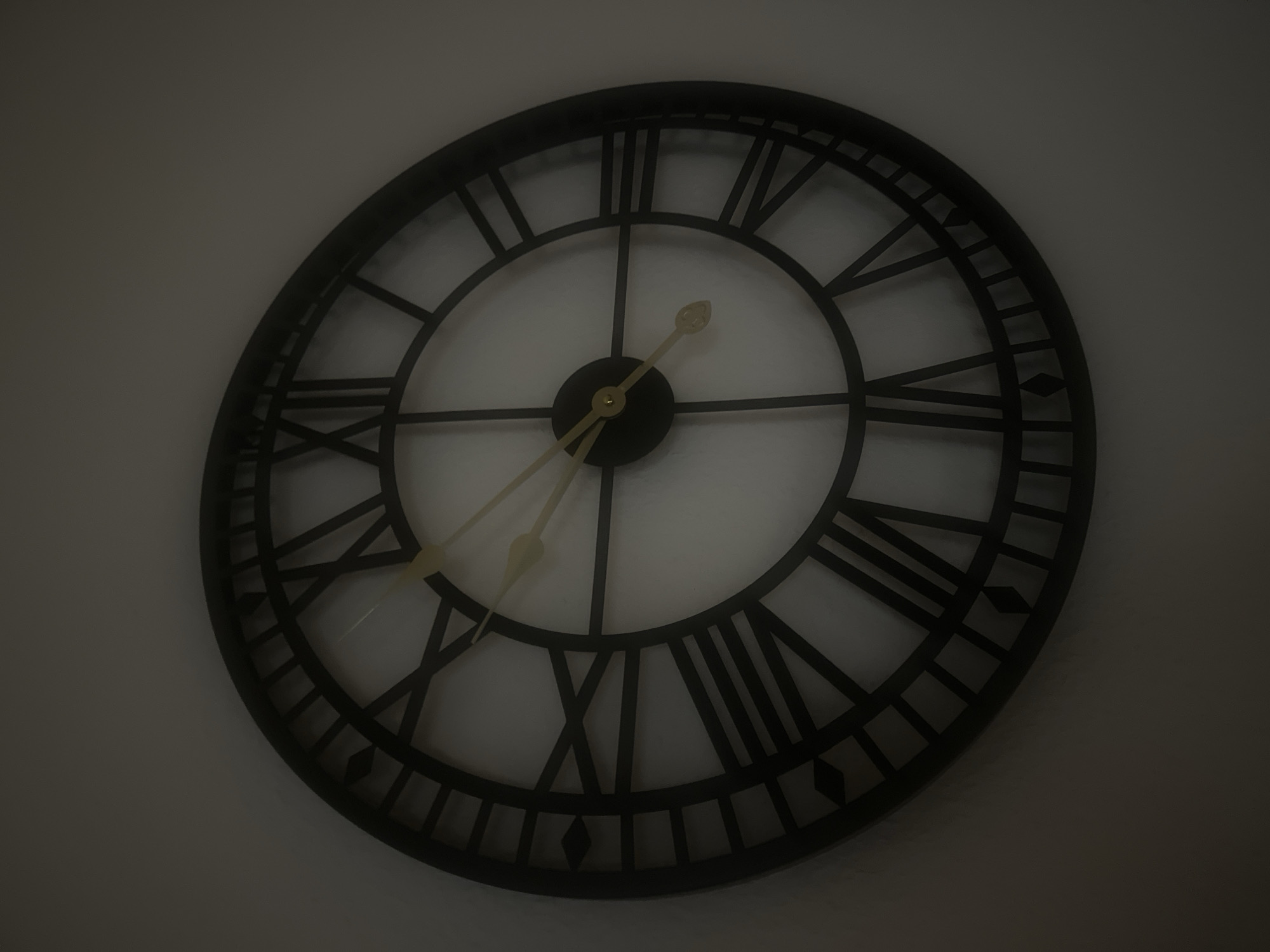 A close-up, slightly angled, indoor photograph shows a black, ornate clock with Roman numerals on a white wall. The clock has a thin black frame and a white face. The hour hand is pointing slightly past the IV (4) and the minute hand is pointing slightly past the V (5). The clock is mounted on a plain white wall. There is no indication of the city of Bautzen, Germany.