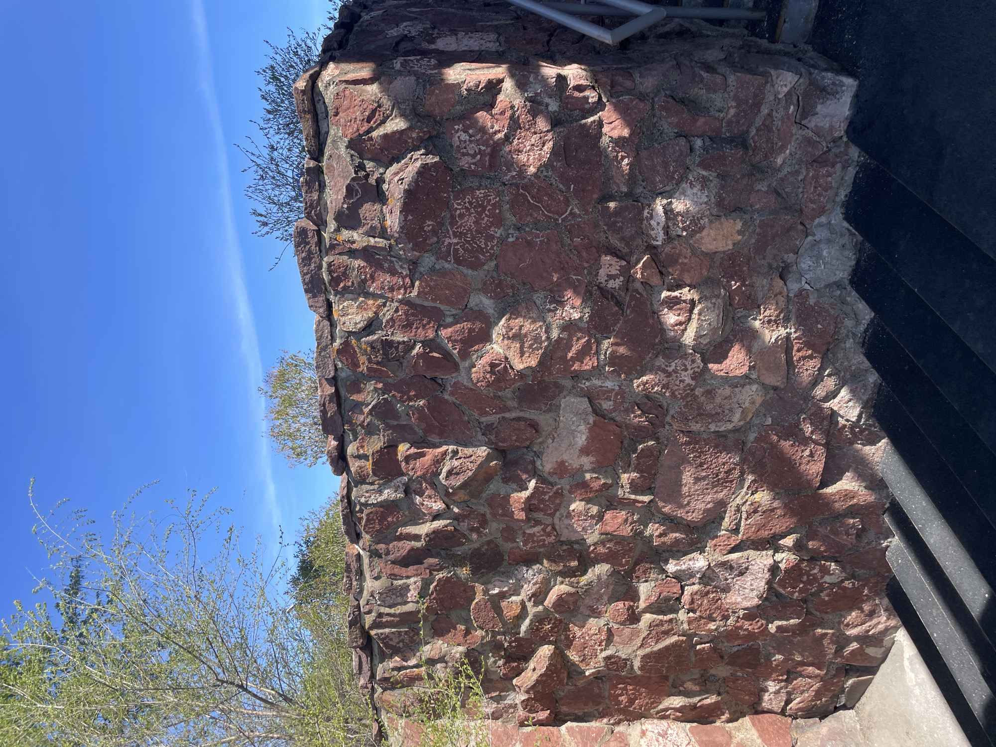 The image shows a portion of the Paraskeva Pyatnitsa Chapel in Krasnoyarsk, Russia. Specifically, it depicts a section of the chapel's exterior wall constructed of reddish-brown stone, likely volcanic rock. The wall is built in a rough, layered fashion, with stones of varying sizes and shapes. Mortar fills the gaps between the stones. Vegetation is visible growing on the edges of the wall, including small plants and branches of trees. In the upper right corner of the frame, a part of a black metal structure (likely steps) is visible, casting a shadow on the stone wall. The background reveals a clear, blue sky. The photograph is taken from a low angle.