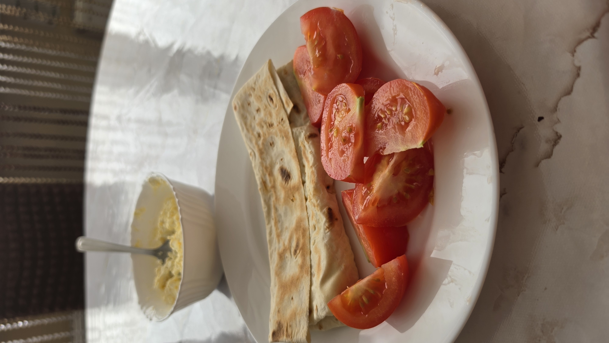 A white plate of food sits on a table. The plate contains two folded flatbreads and several slices of chopped tomatoes. In the background, out of focus, is a white bowl containing what appears to be scrambled eggs or mashed potatoes, with a fork resting in it. The table has a light-colored, textured surface.