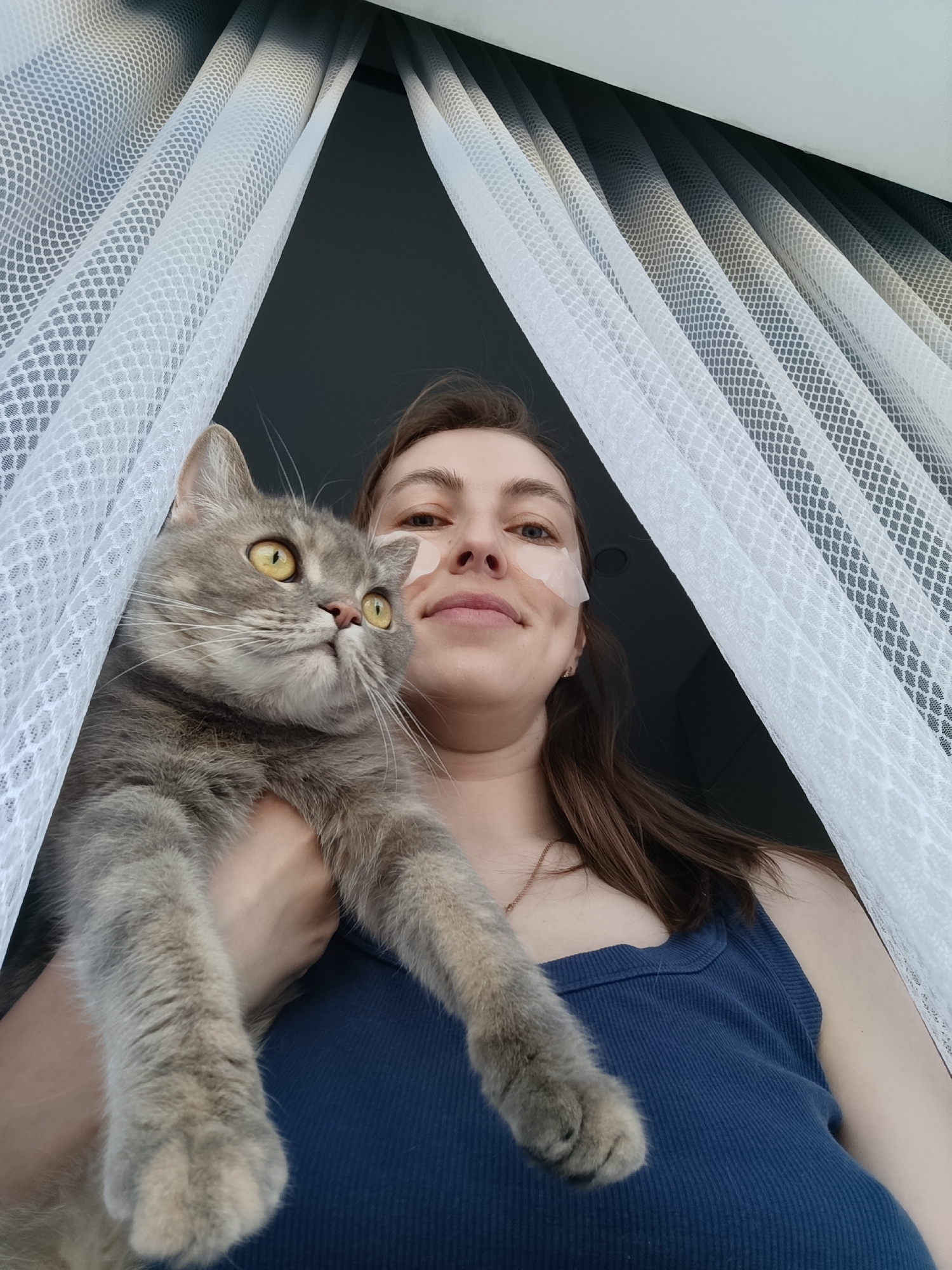 A woman with light skin, wearing a blue tank top and white adhesive patches on her face, is holding a gray tabby cat. The cat has yellow eyes and is looking up and to the left. They are positioned near a window covered with white, mesh-like curtains. The location is Chaikovsky, Russia.