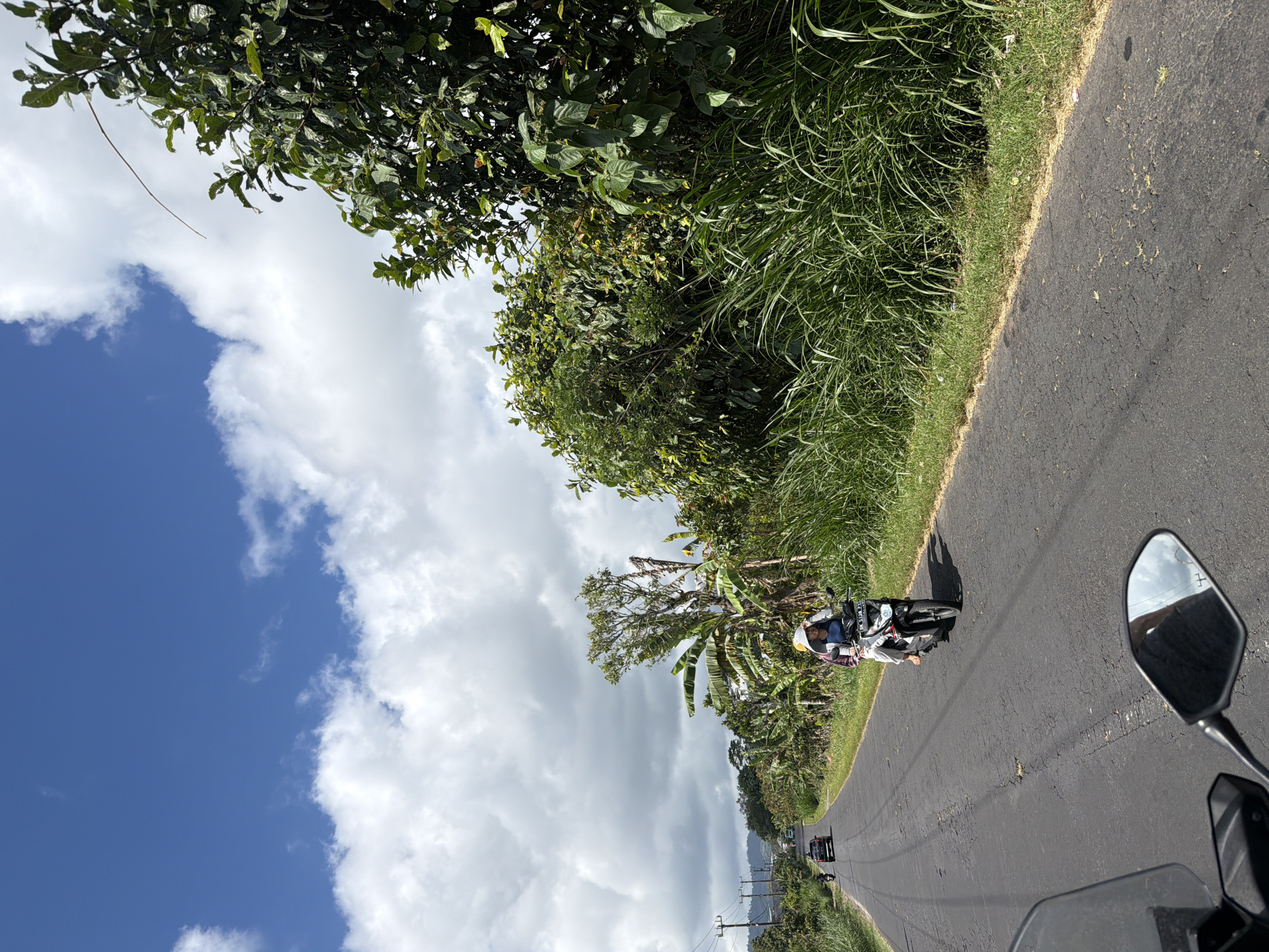A person rides a scooter on an asphalt road lined with lush green vegetation and trees. The sky above is a vibrant blue with scattered white clouds. In the distance, a car is visible, also traveling on the road. The scene appears to be in a rural or semi-rural area, possibly in Kintamani, Indonesia, given the tropical flora and the overcast yet sunny conditions. The perspective of the image is from another scooter, showing its side mirror in the foreground.