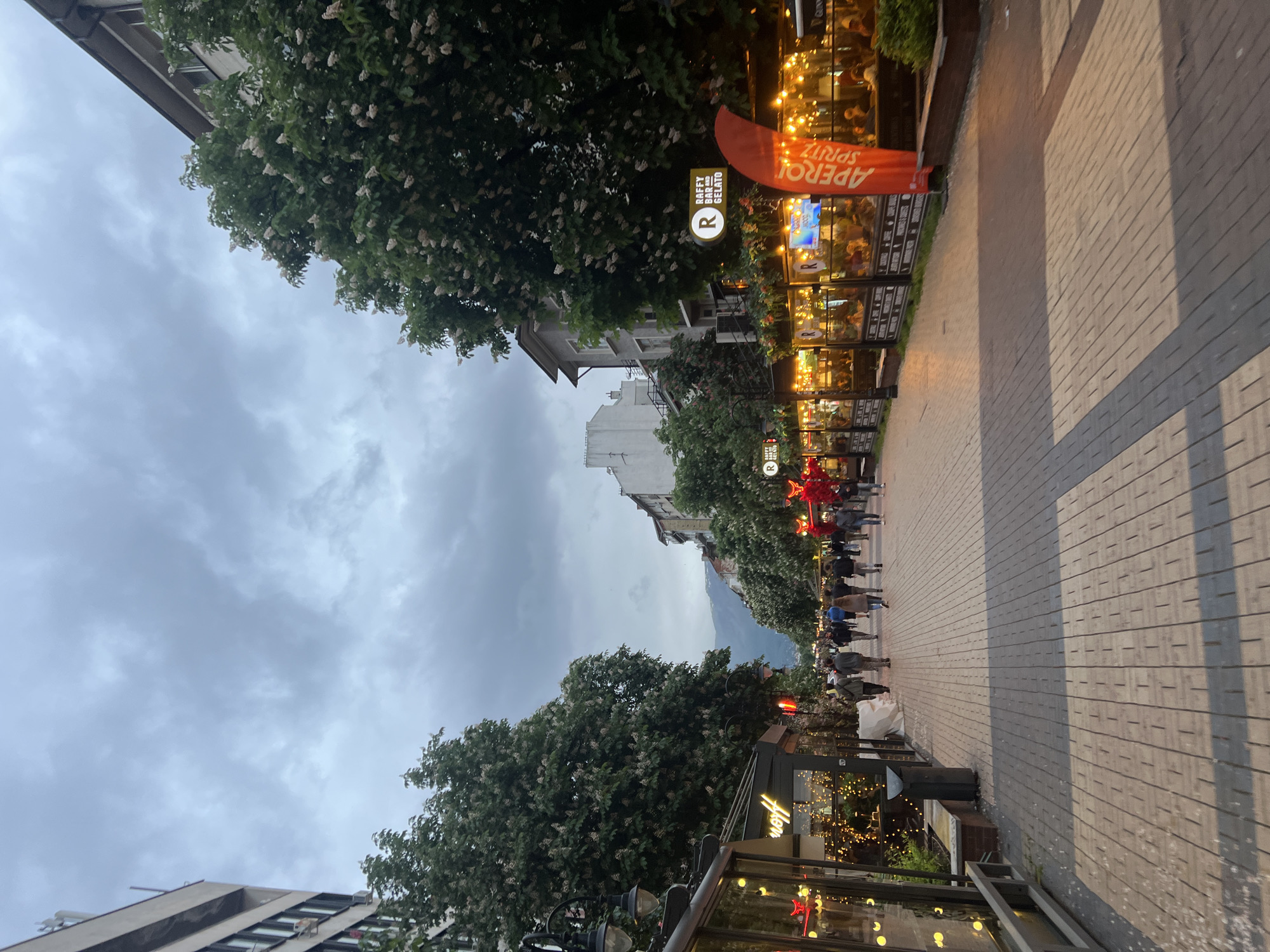 The image depicts a street scene in Sofia, Bulgaria, along Vitosha Boulevard. The perspective is looking down the pedestrianized walkway. On the right are outdoor dining areas with string lights and an "Aperol Spritz" advertisement; the Raffy Bar/Gelato sign is also visible. People are walking along the boulevard. Above, trees partially obscure the cloudy sky and buildings. In the background is a view of Vitosha Mountain. The street is paved with rectangular tiles.